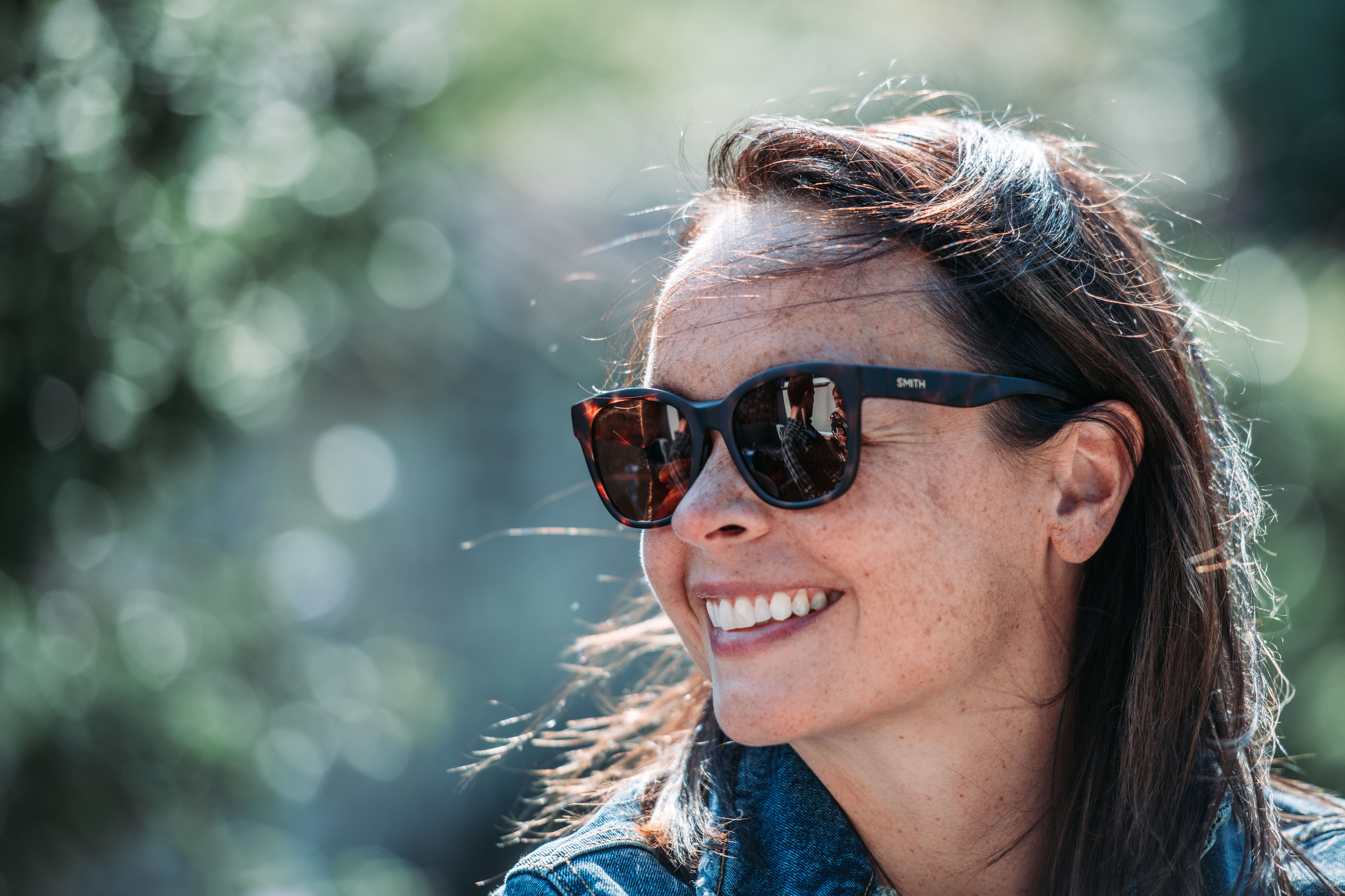 A woman wearing sunglasses and a denim jacket smiles while looking off to the side.