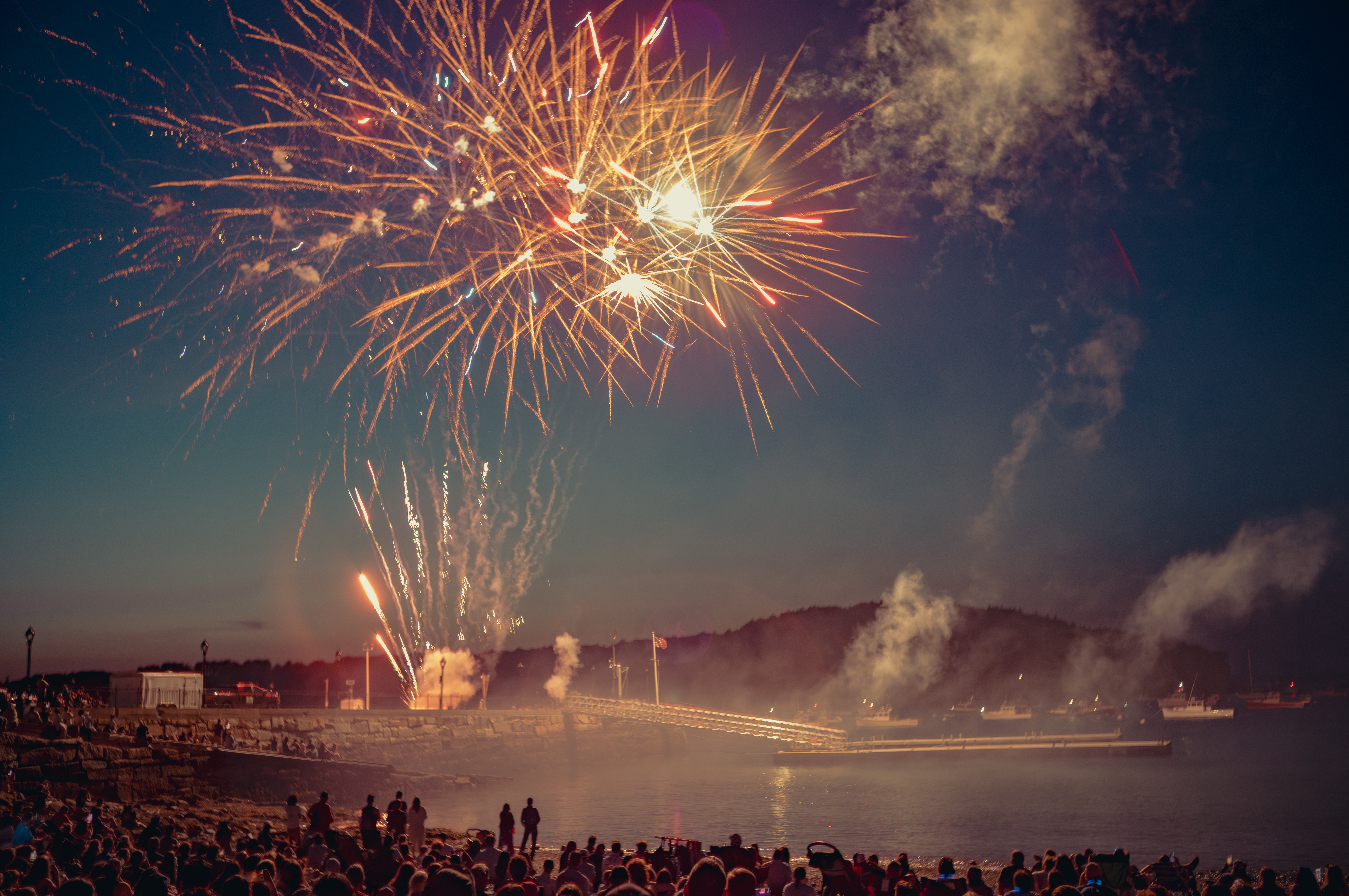 Alt text: Fireworks over the water in Bar Harbor, Maine, with boats in the harbor and a packed beach watching from below.