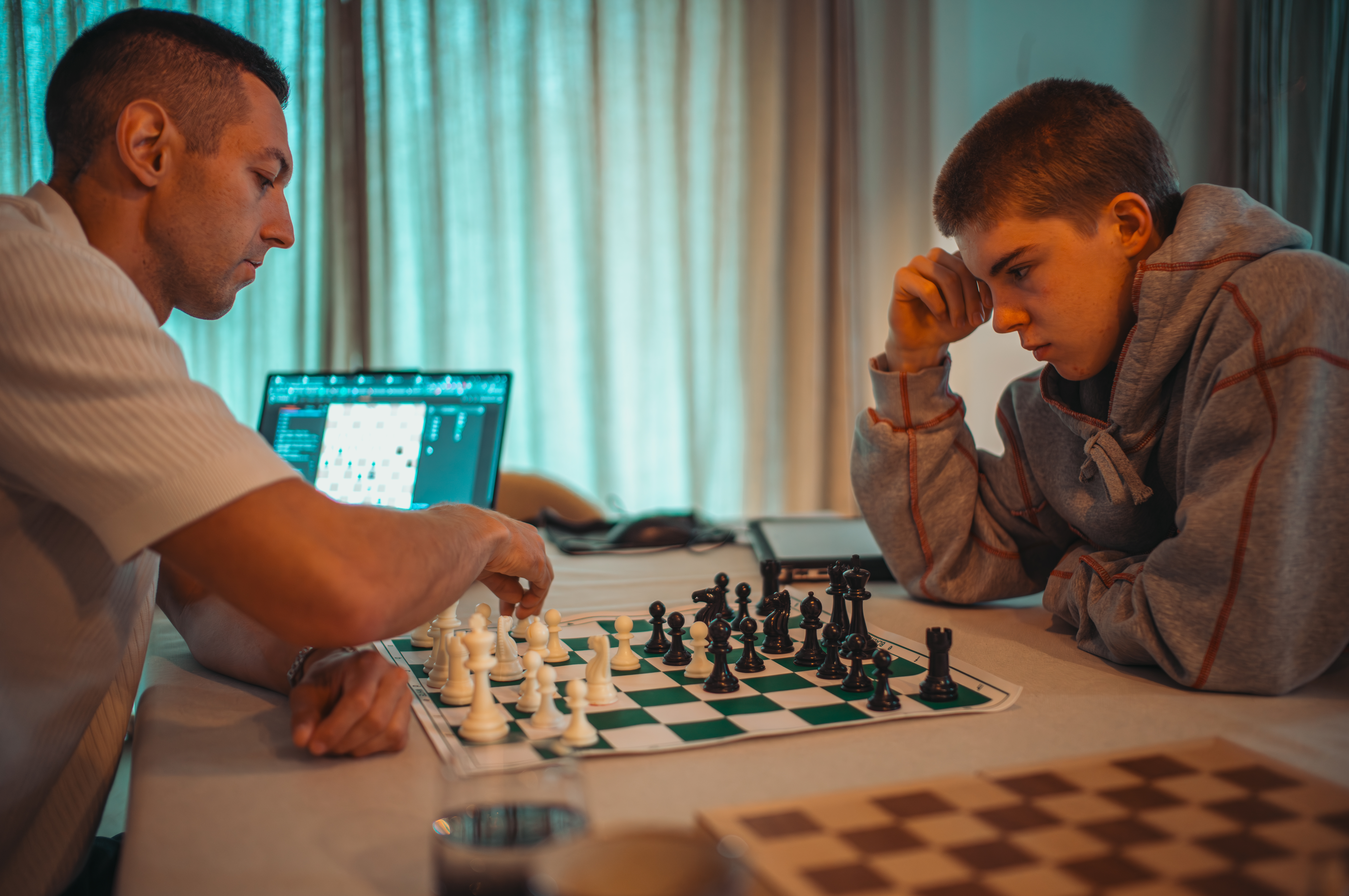 A man and a boy play chess, the boy leaning forward in concentration as the man moves a piece.
