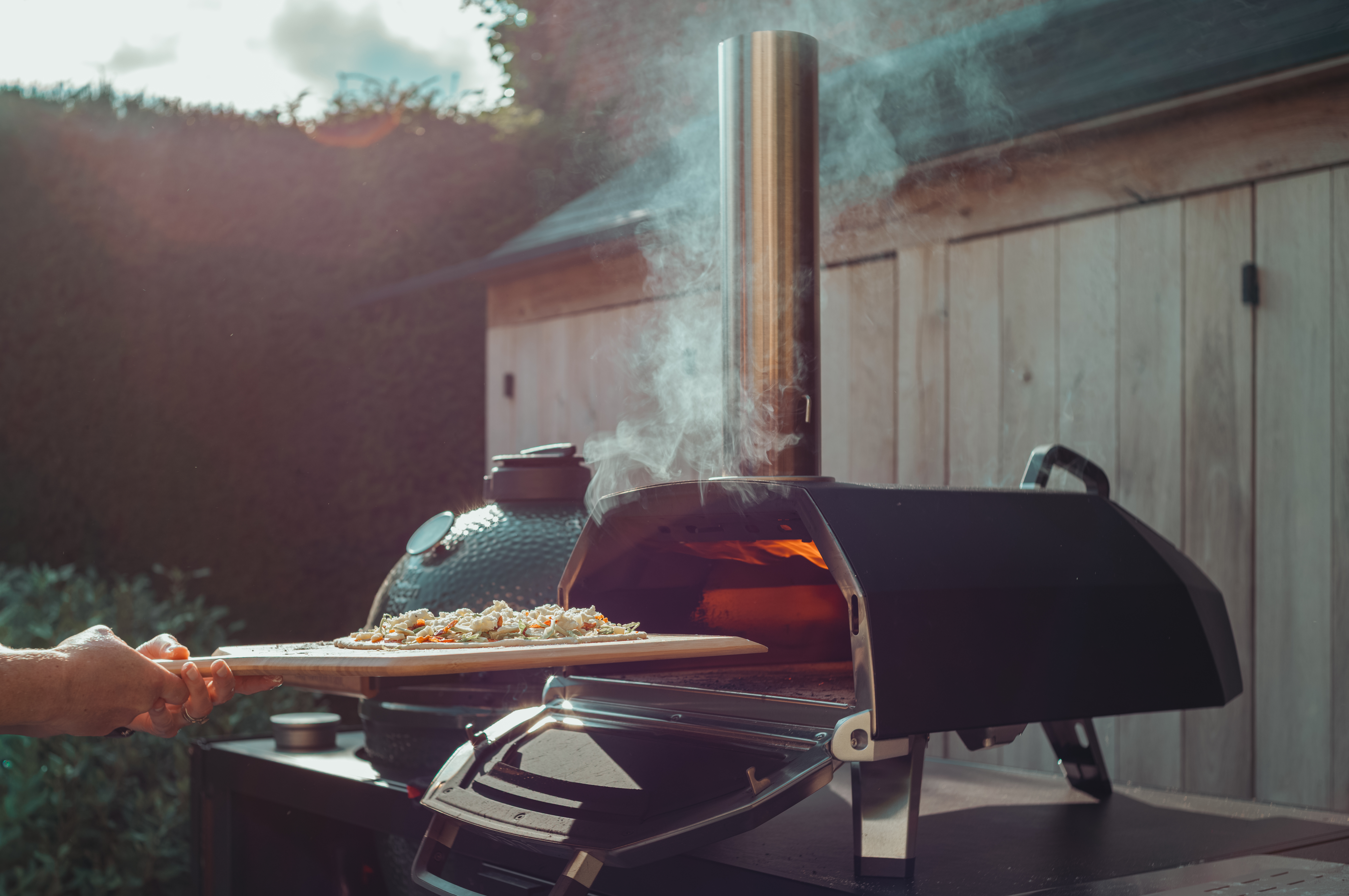 A homemade pizza is being slid into a smoking outdoor pizza oven.