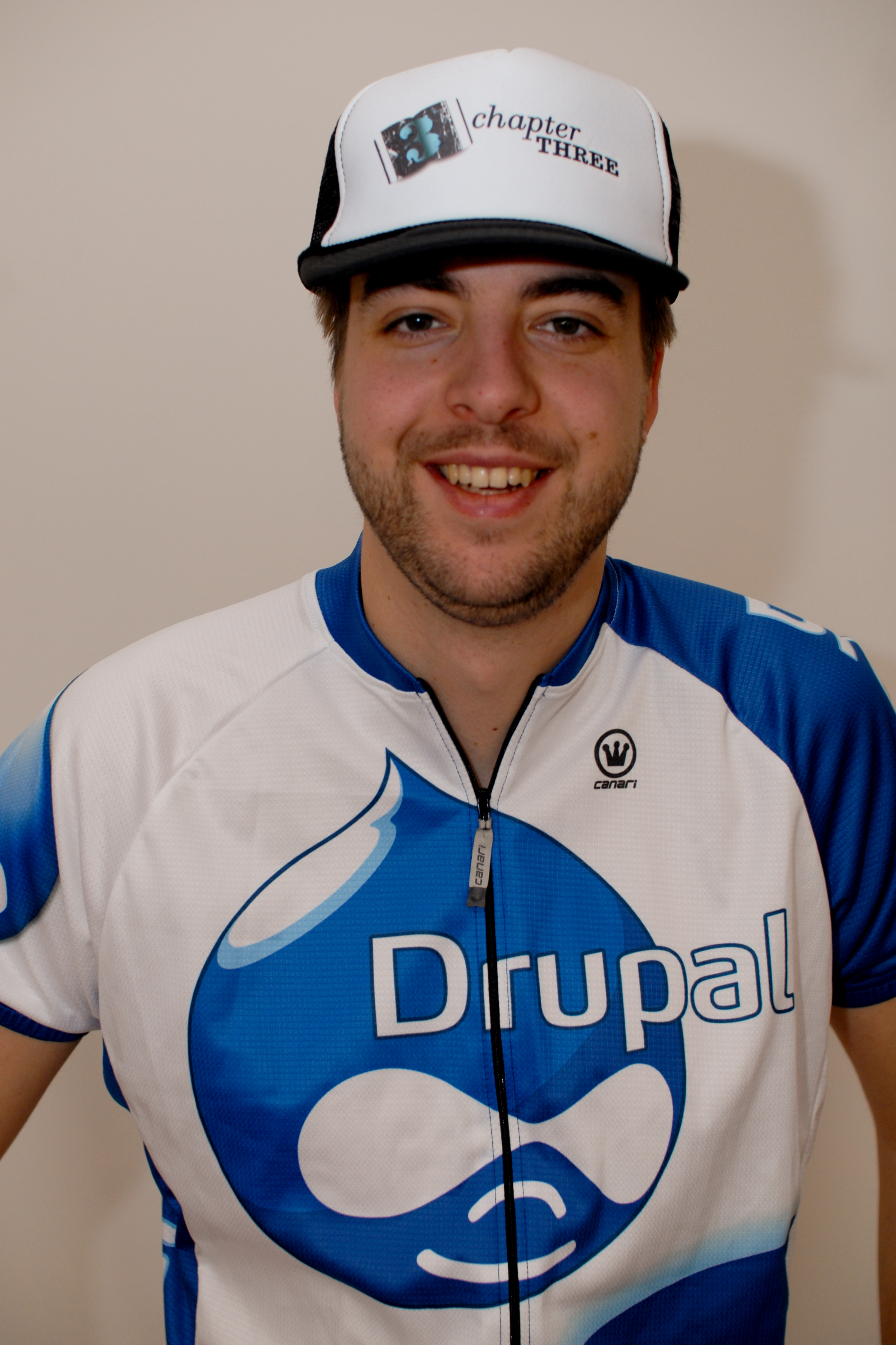 A smiling person wears a Drupal cycling jersey and a cap with "Chapter Three" printed on it.
