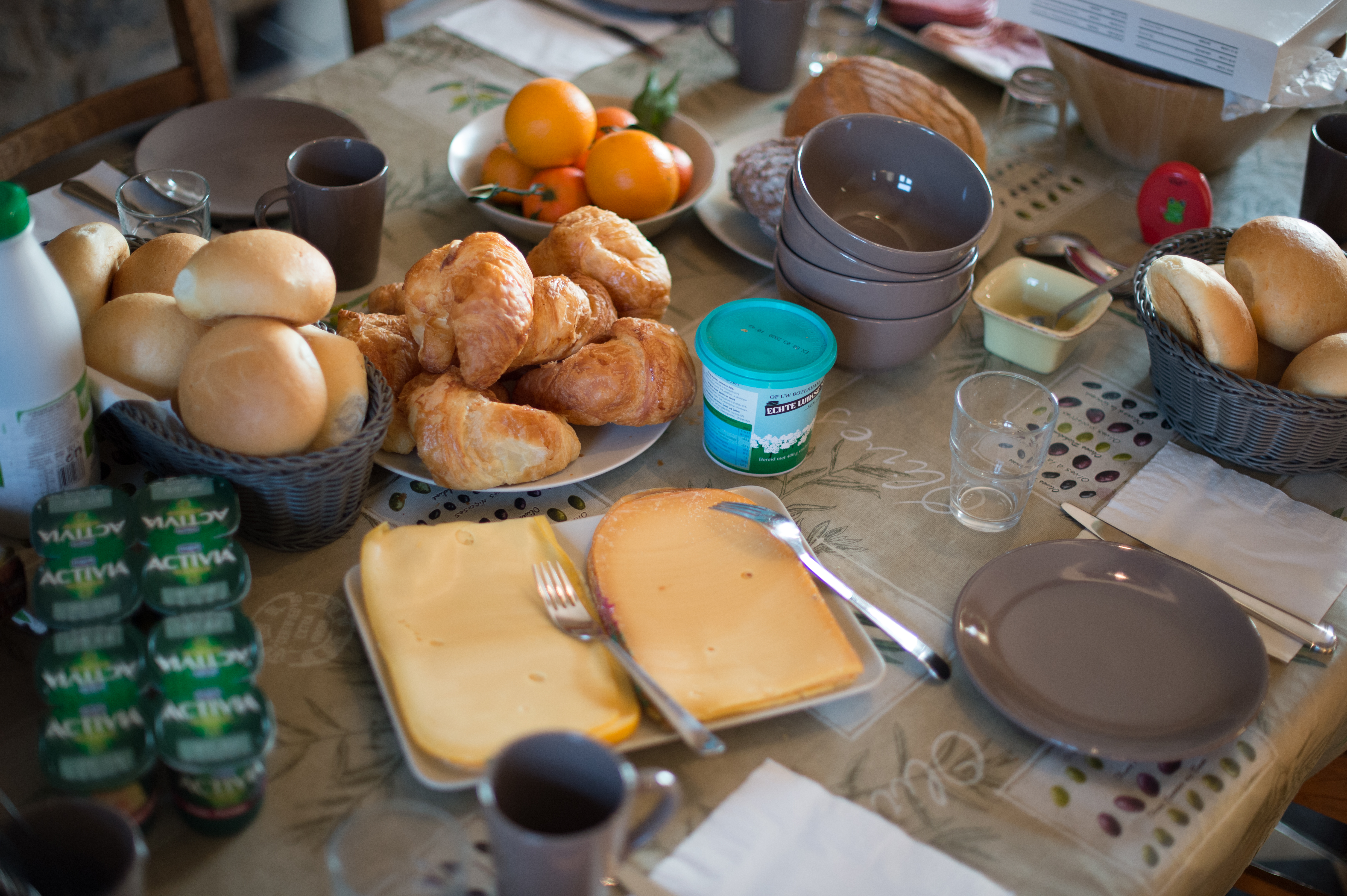 A breakfast table with croissants, bread rolls, cheese, yogurt, fruit, and drinks, set for multiple people.