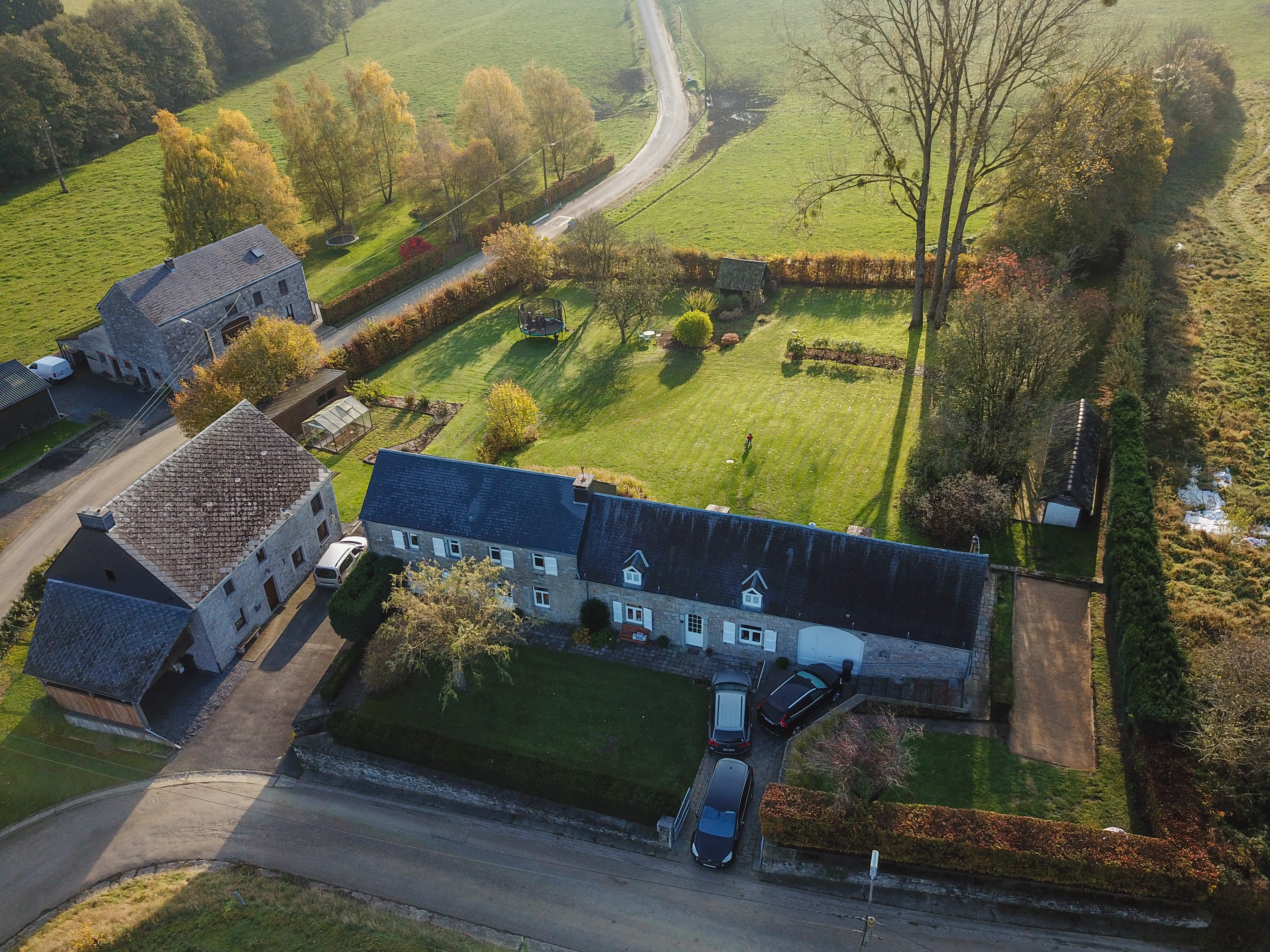 Aerial view of a countryside house with a large garden, parked cars, and a person standing on the lawn.