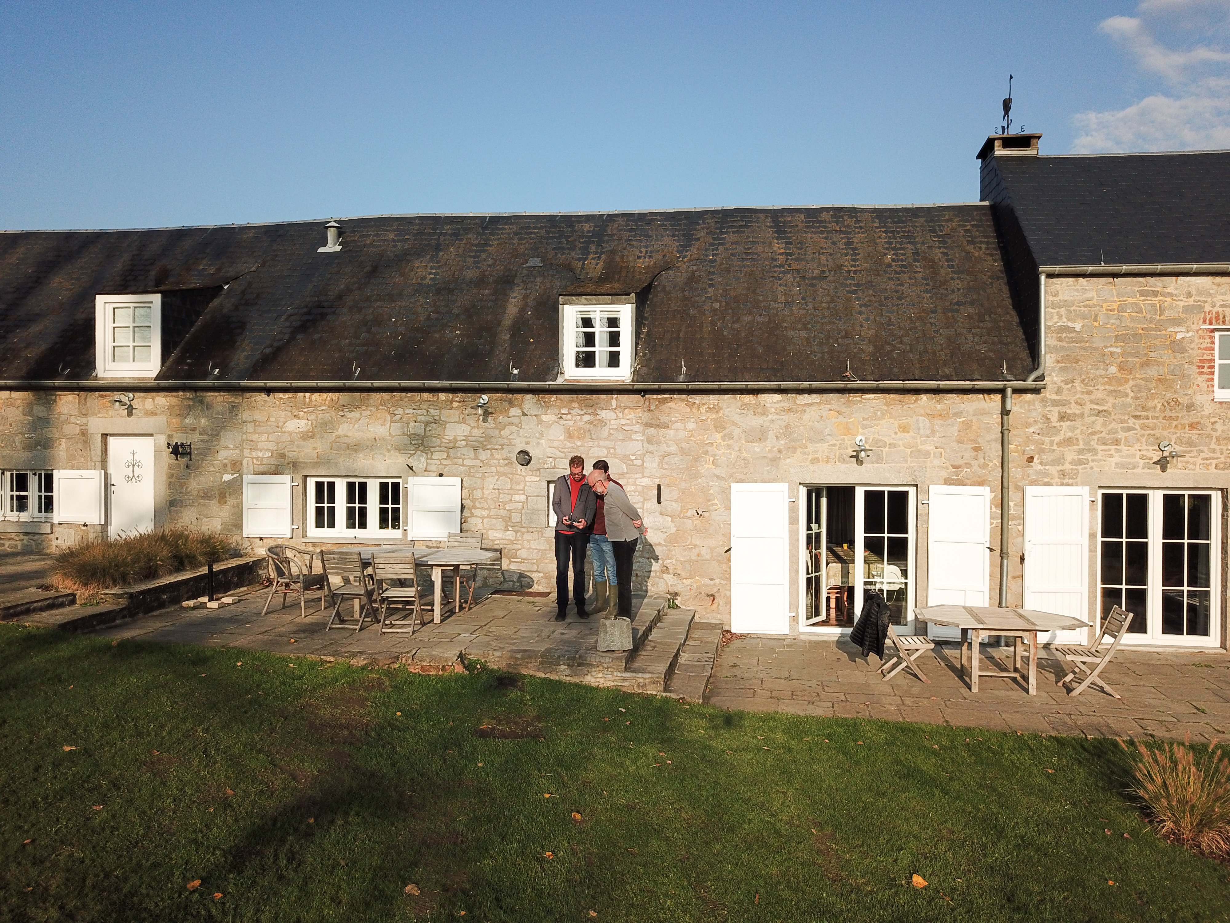 Two people stand on a stone patio, operating a drone in front of a rustic house with white shutters.