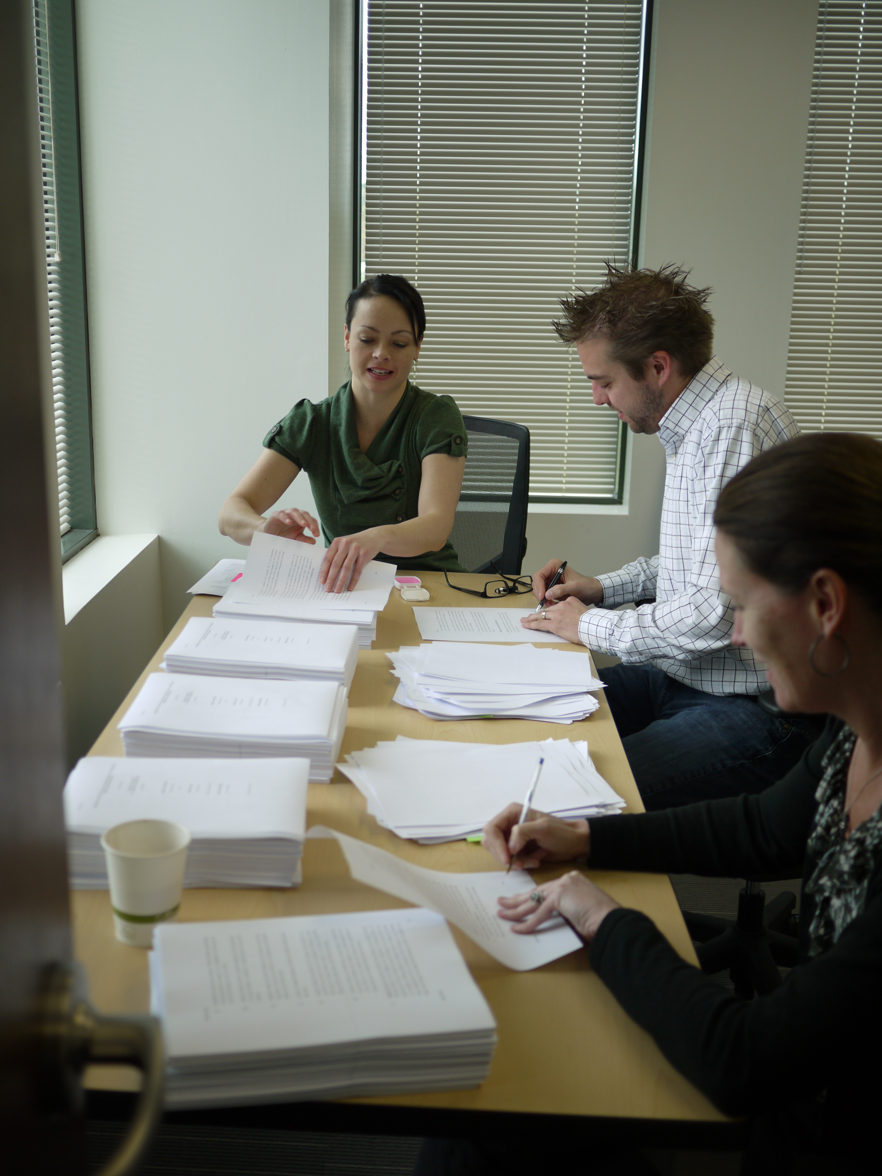 Three people sit at a table sorting and signing documents, with stacks of paper around them.