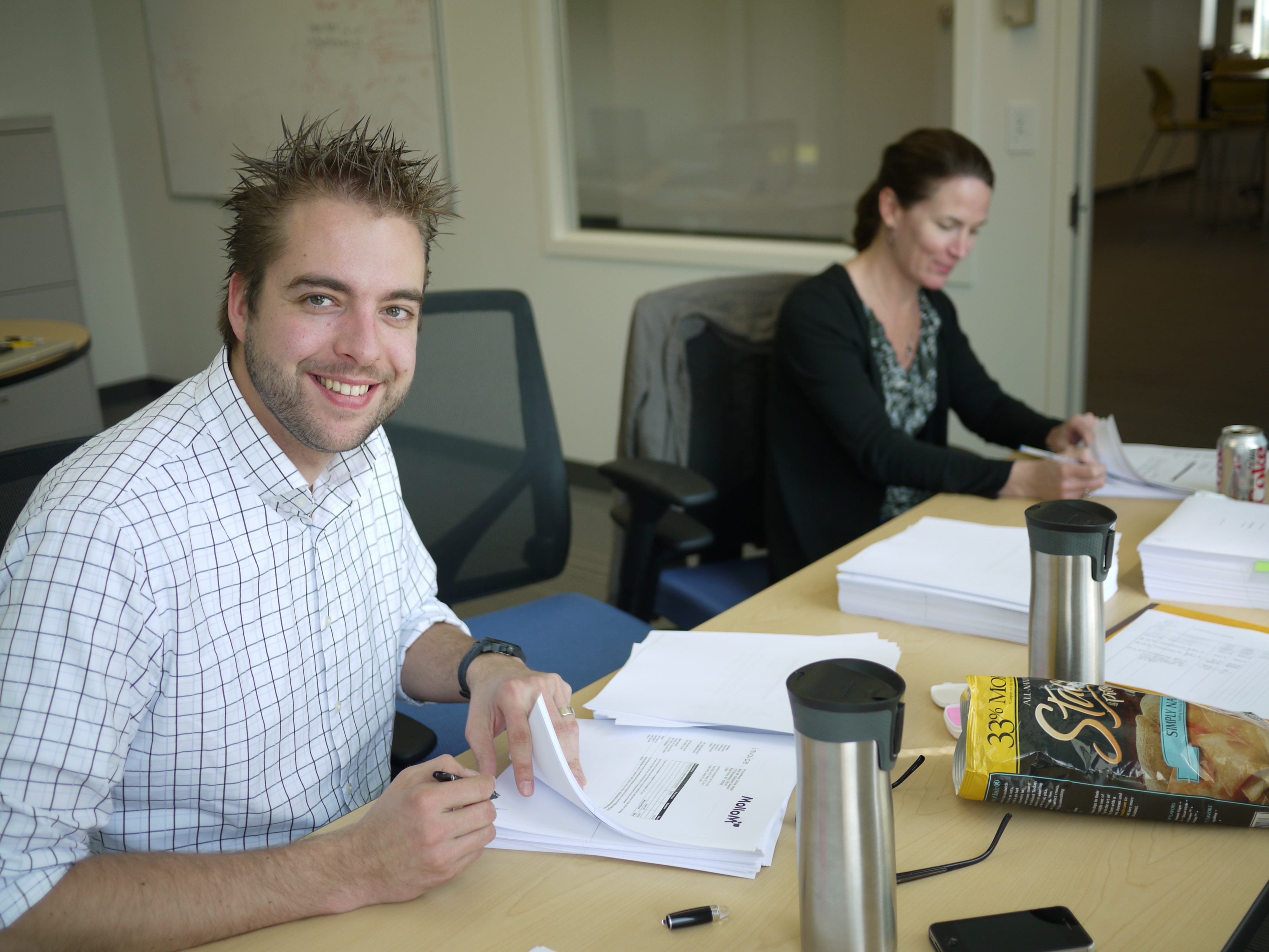 Two people sit at a table signing documents, with stacks of papers, coffee mugs, and snacks nearby.