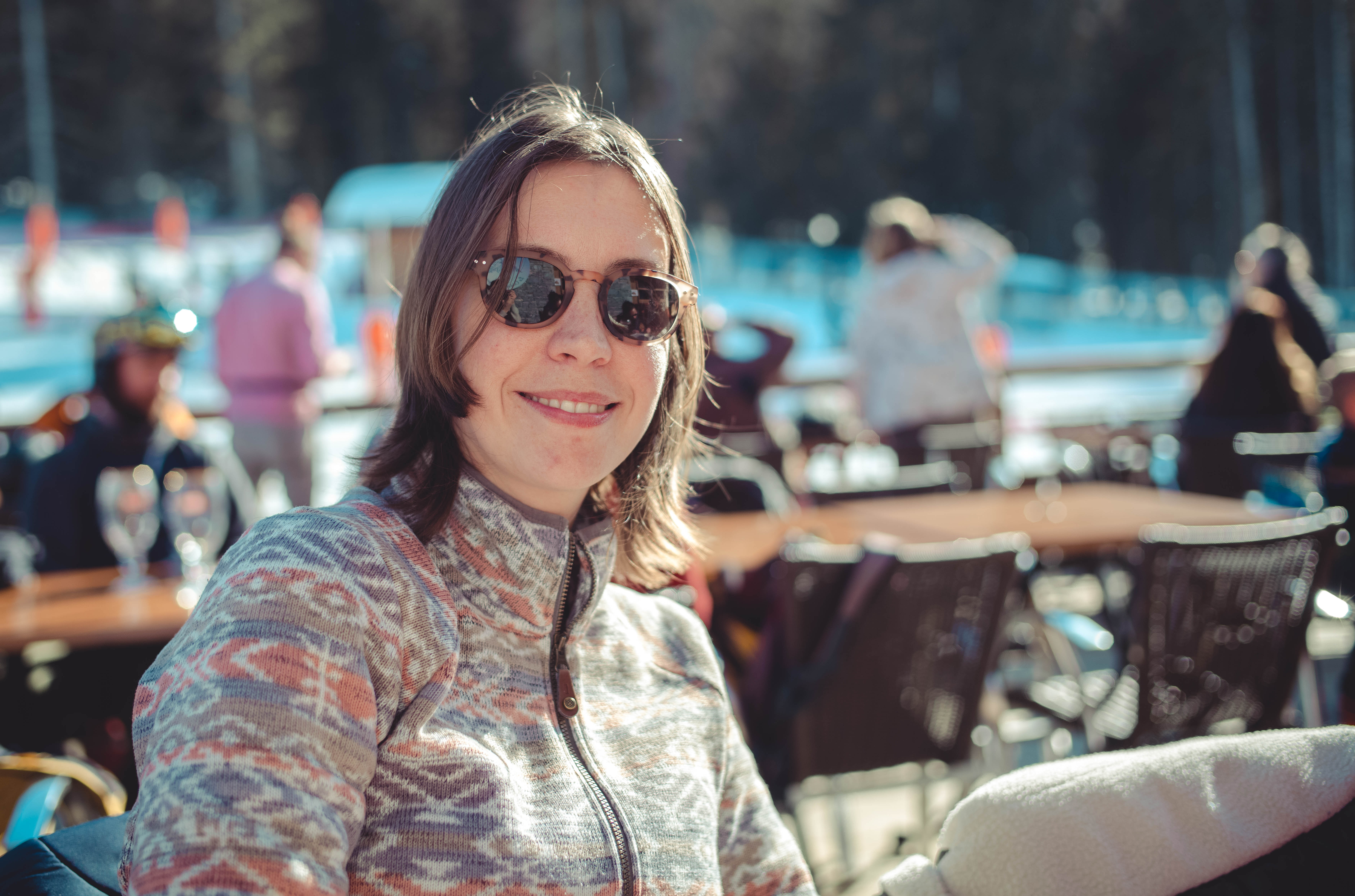 A woman wearing sunglasses and a patterned jacket smiles while sitting at an outdoor seating area.