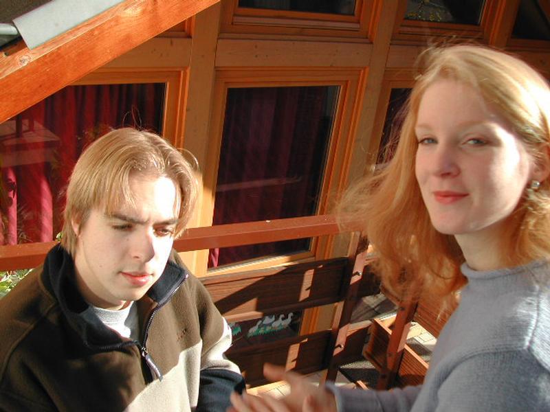 A young man and woman sit on a wooden balcony, with the woman smiling slightly and looking at the camera.