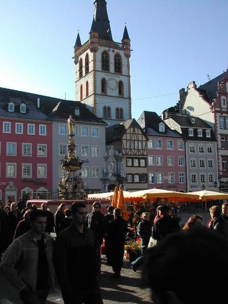 A busy market square with people walking, colorful buildings, a tall church tower, and a decorative fountain.