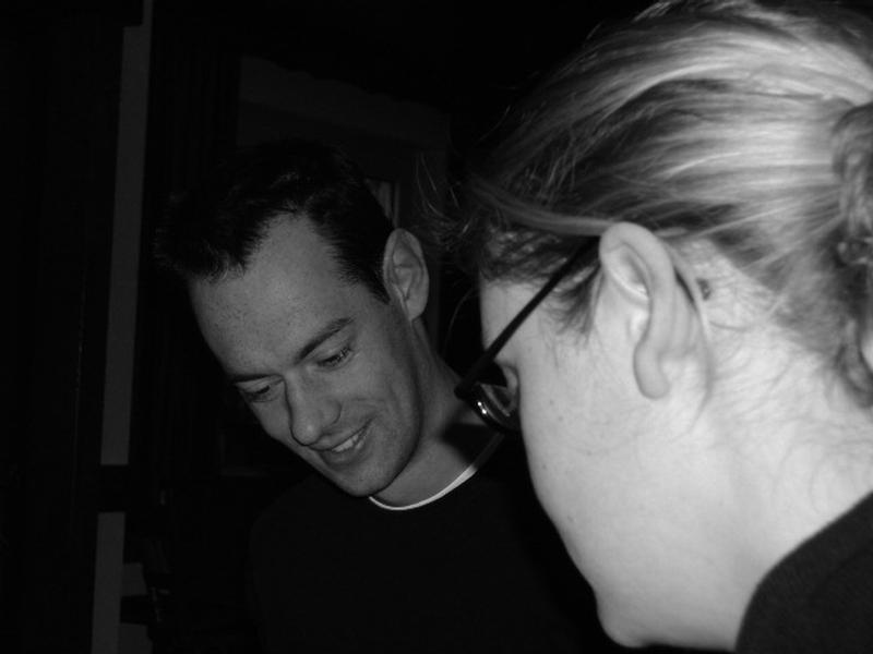 A man and a woman are engaged in conversation, with the man smiling and looking downward.