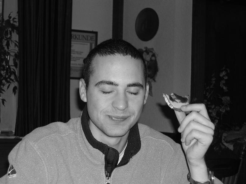 A man with closed eyes smiles while holding a piece of bread with spread in his hand.