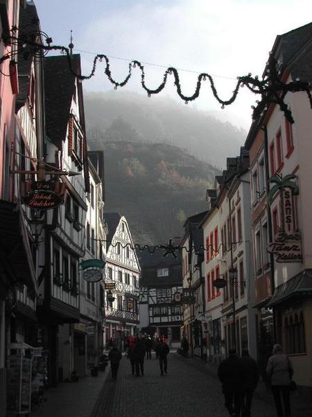 A cobblestone street in a European town with people walking, framed by traditional buildings and festive decorations.