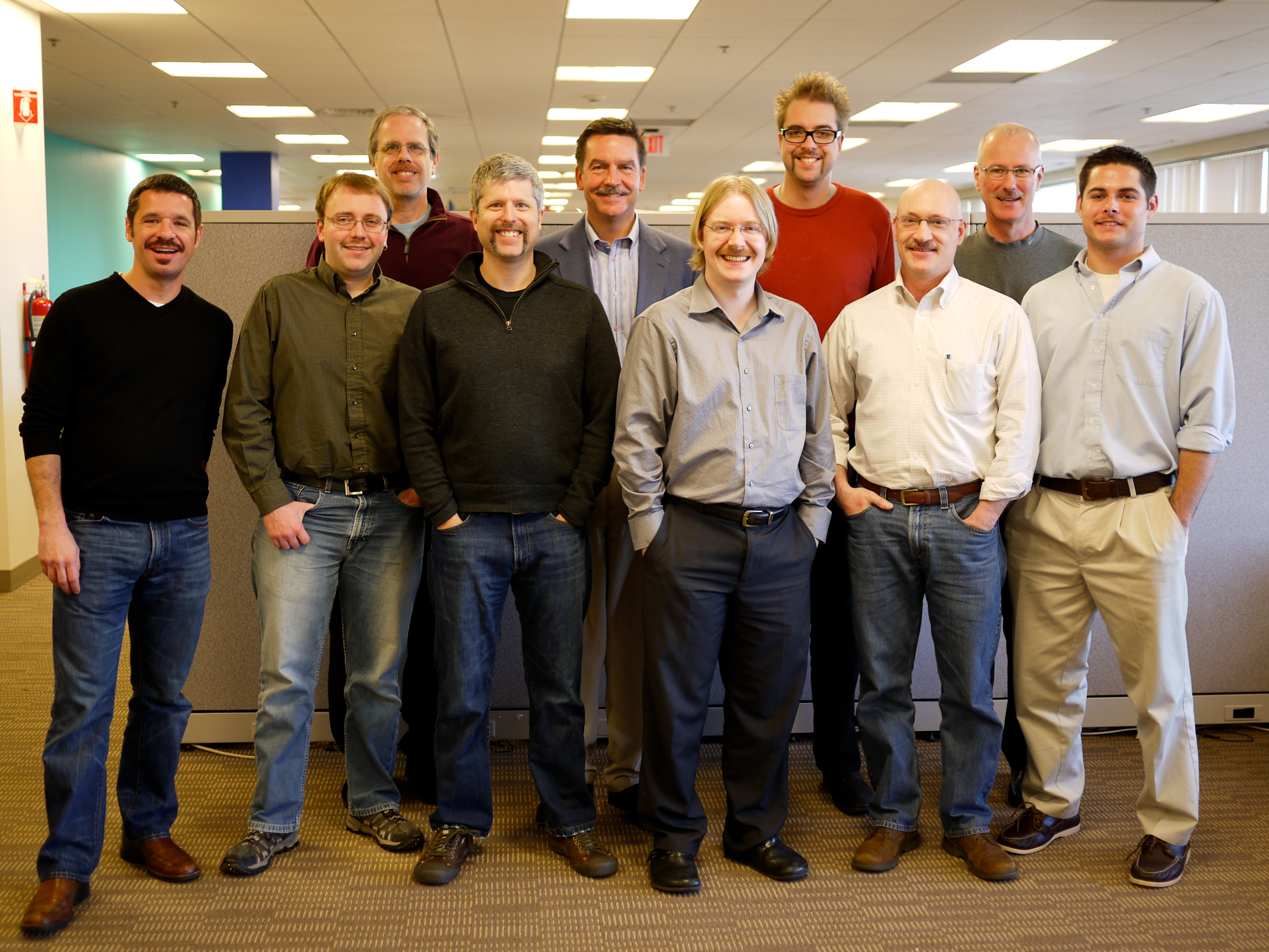 A group of ten men with mustaches stand together in an office, smiling at the camera for Movember.