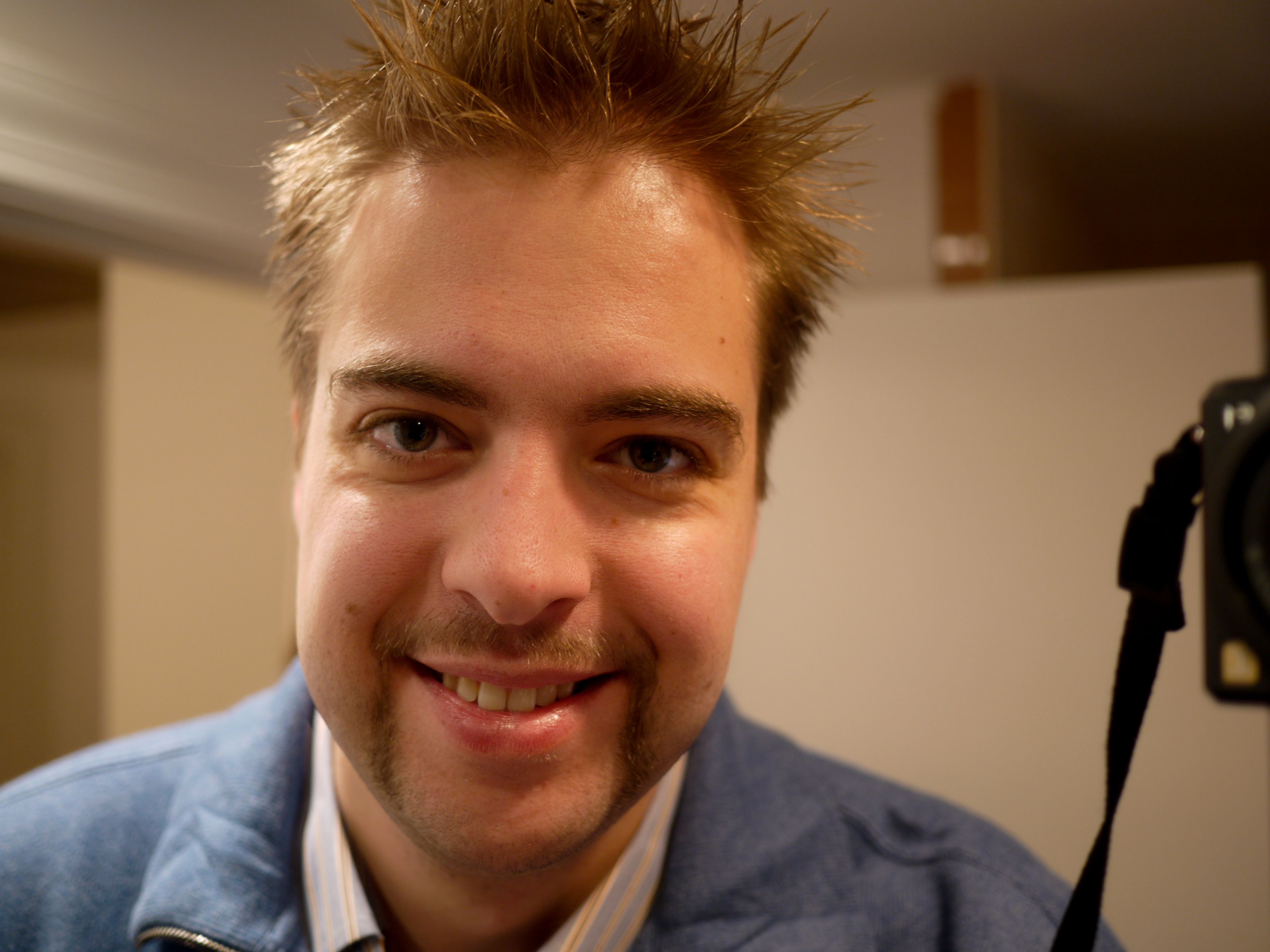 A man with short, spiky hair and a mustache smiles at the camera, wearing a blue jacket indoors.