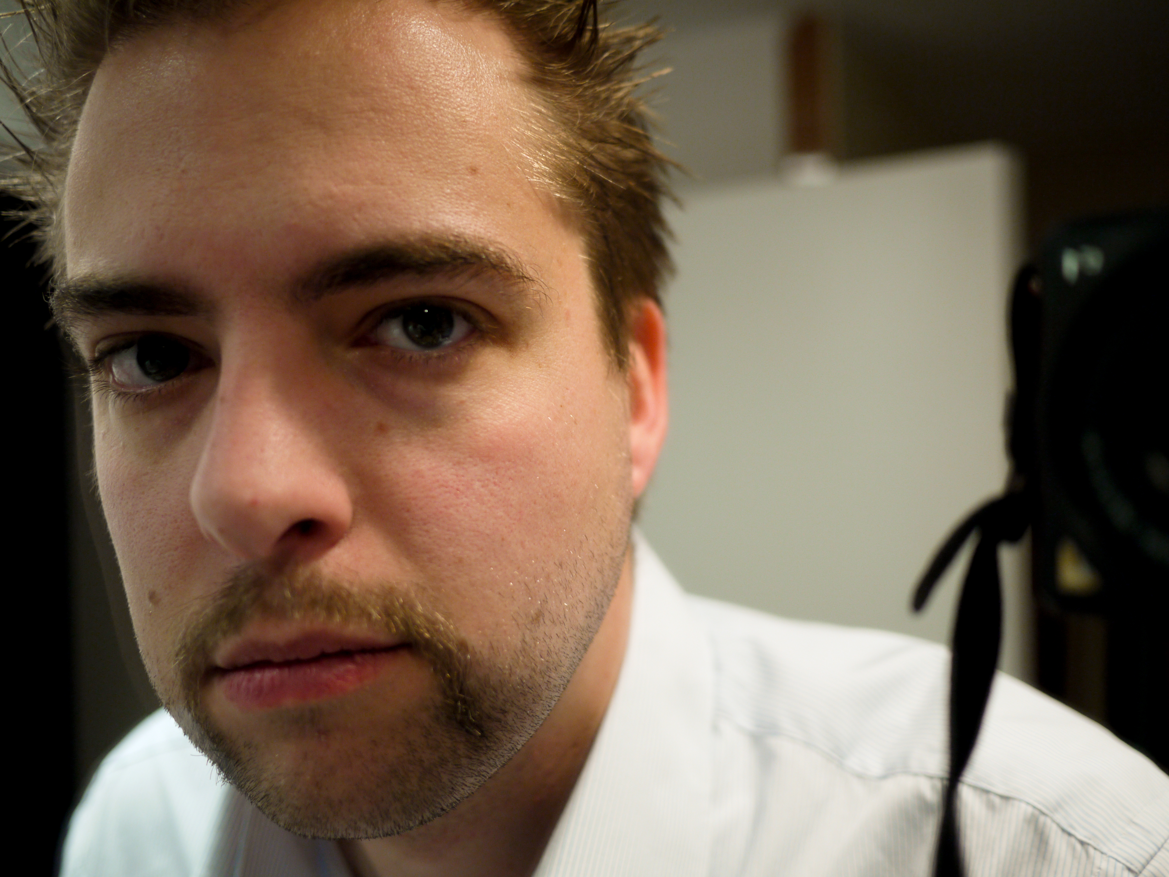 Close-up of a man with a mustache looking directly at the camera, wearing a white shirt indoors.