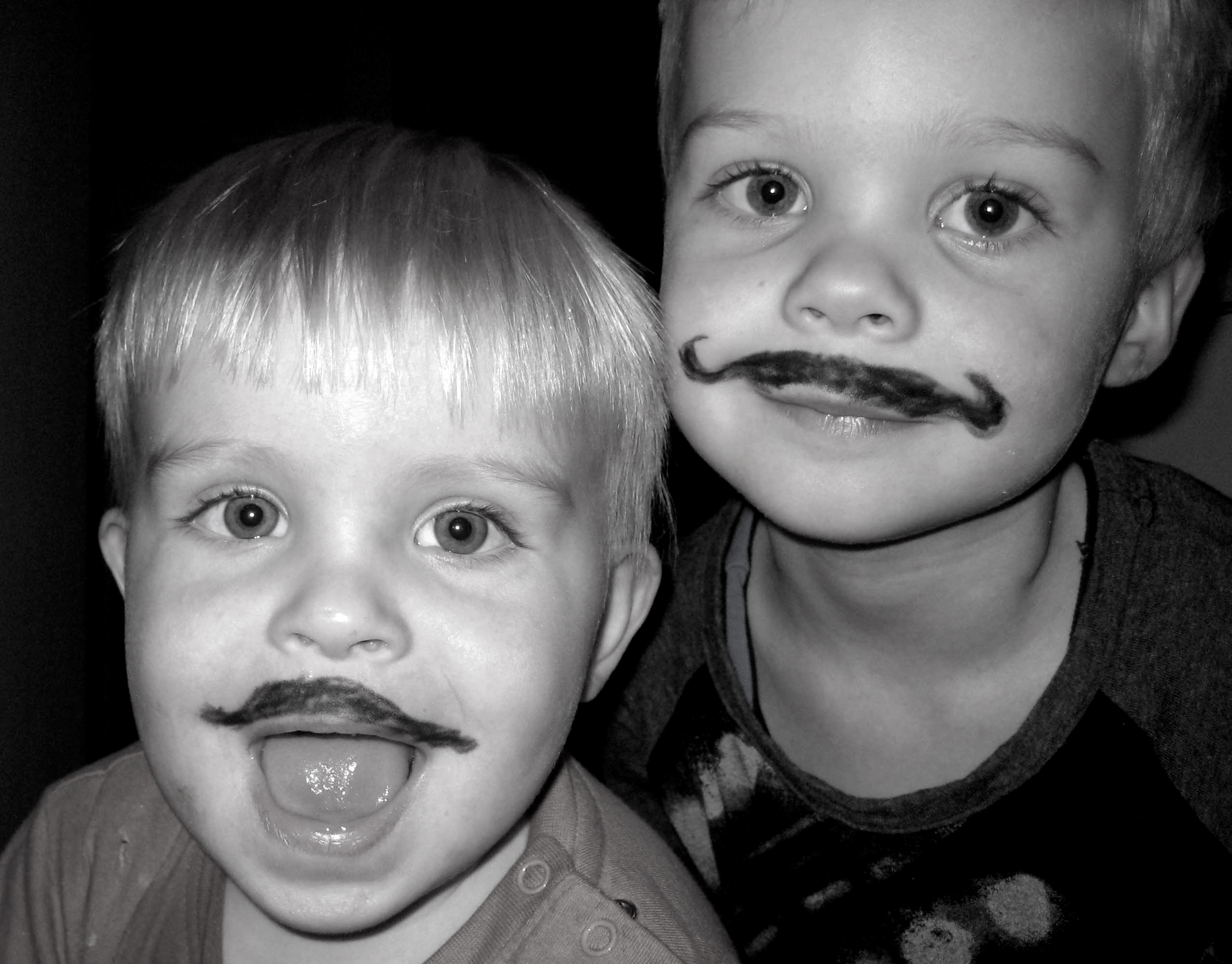 Two young children with drawn-on mustaches smile at the camera, one with an open mouth and the other with a grin.