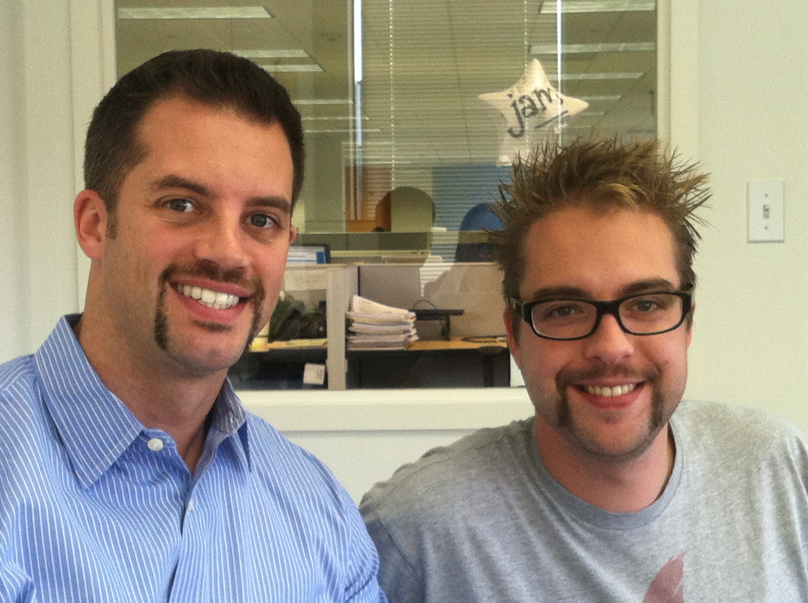 Two men with mustaches smile at the camera in an office setting during Movember 2011.
