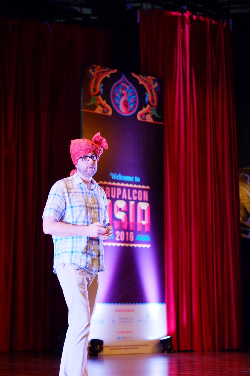 A man wearing a red turban and glasses speaks on stage at DrupalCon Asia 2016, holding a remote.
