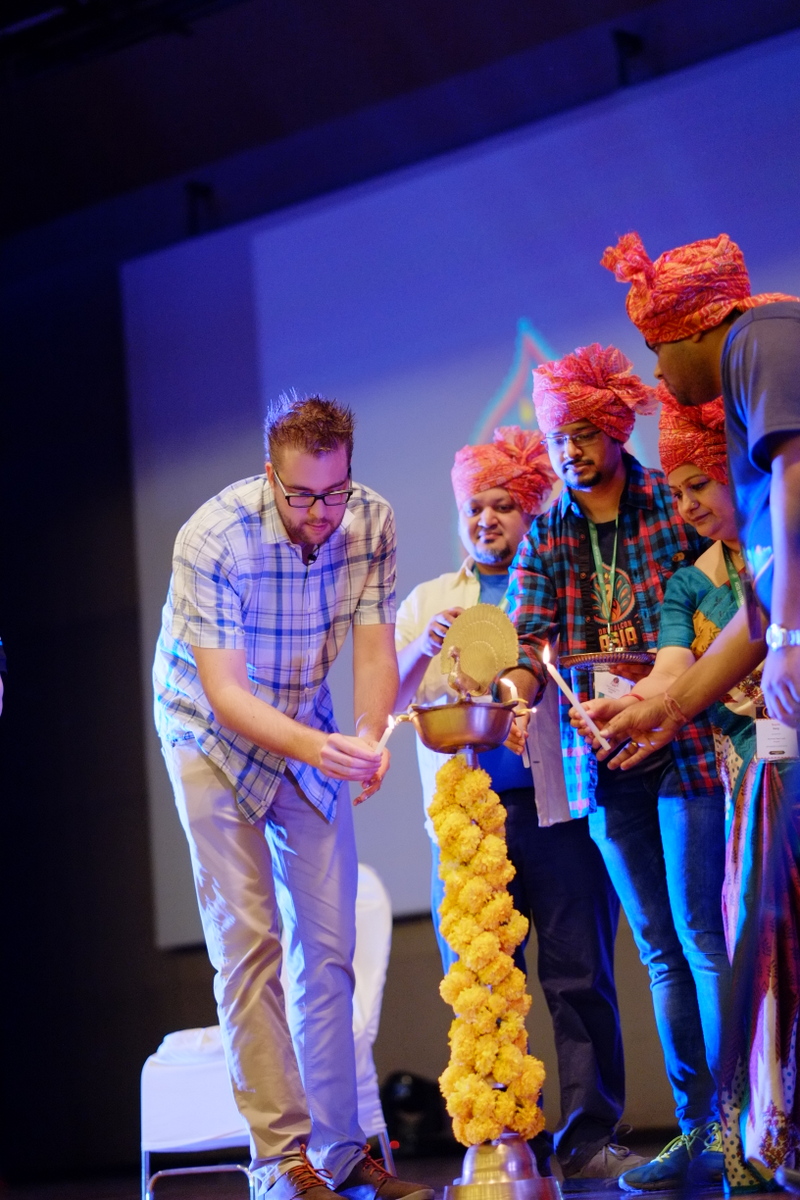A group of people lights candles from a ceremonial lamp at an event, with one person bending forward.