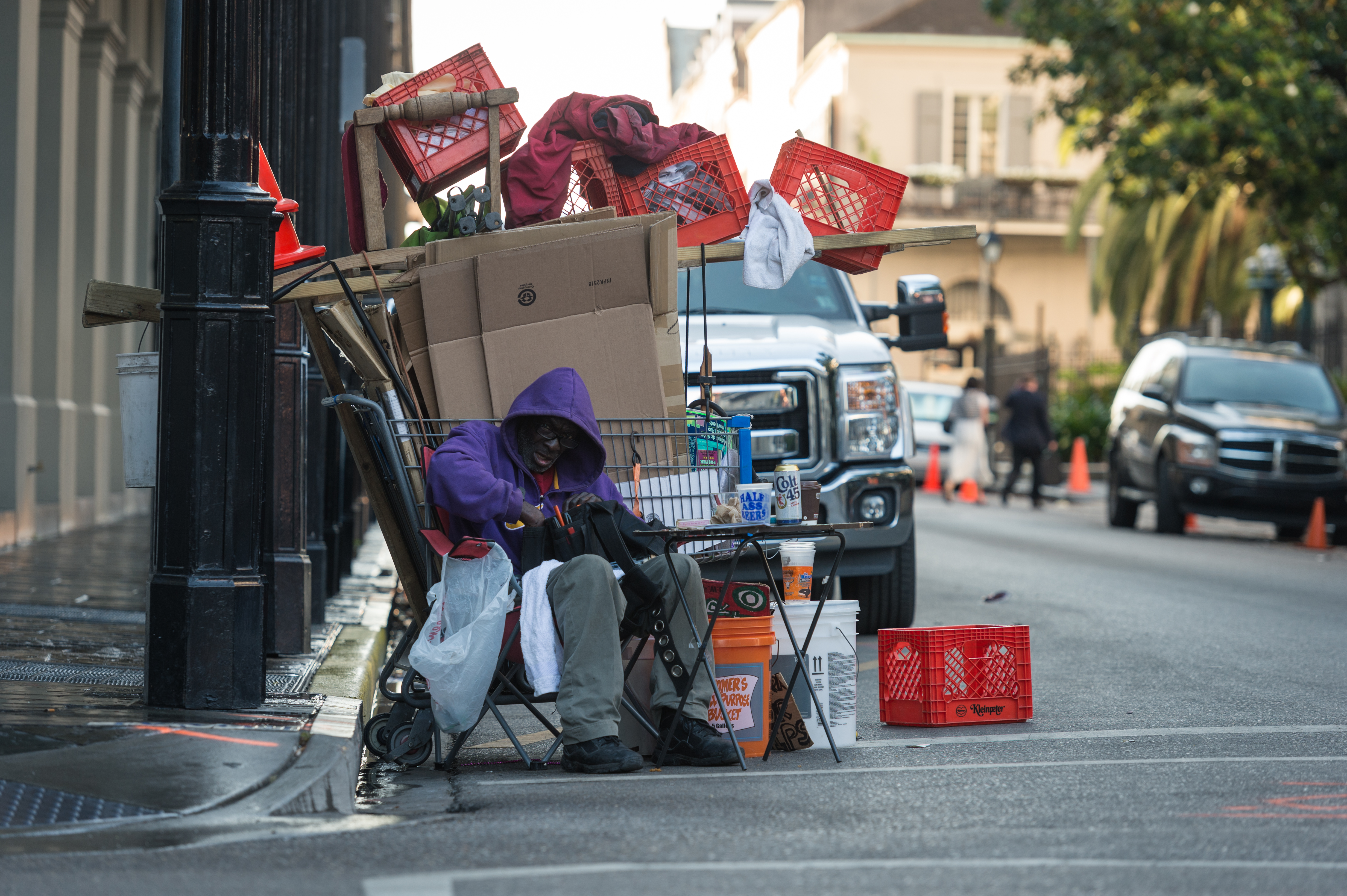 A person in a purple hoodie sits on a chair beside a cart filled with belongings on a city sidewalk.