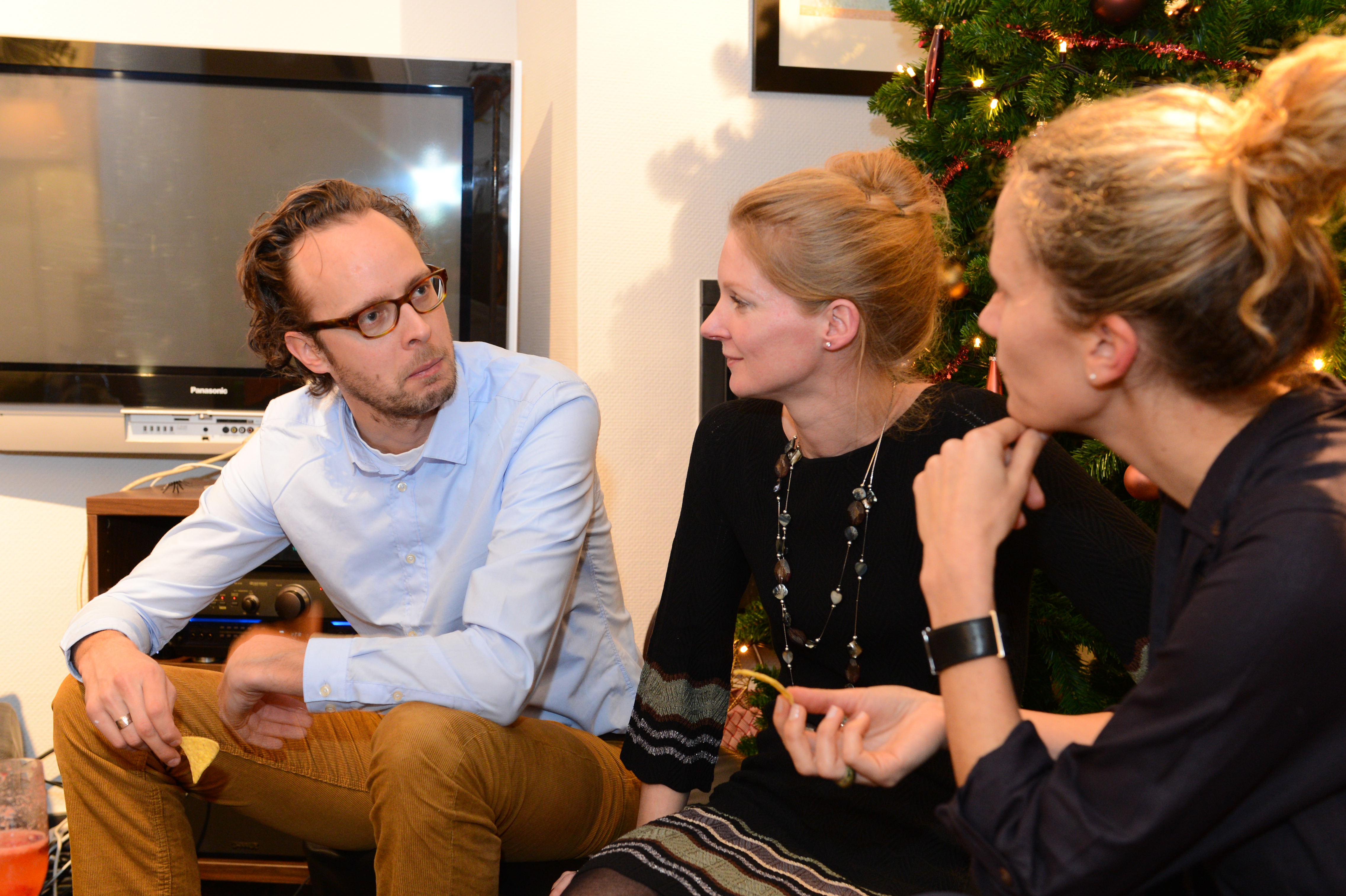 Three people sit near a Christmas tree, engaged in conversation at a New Year's Eve party.