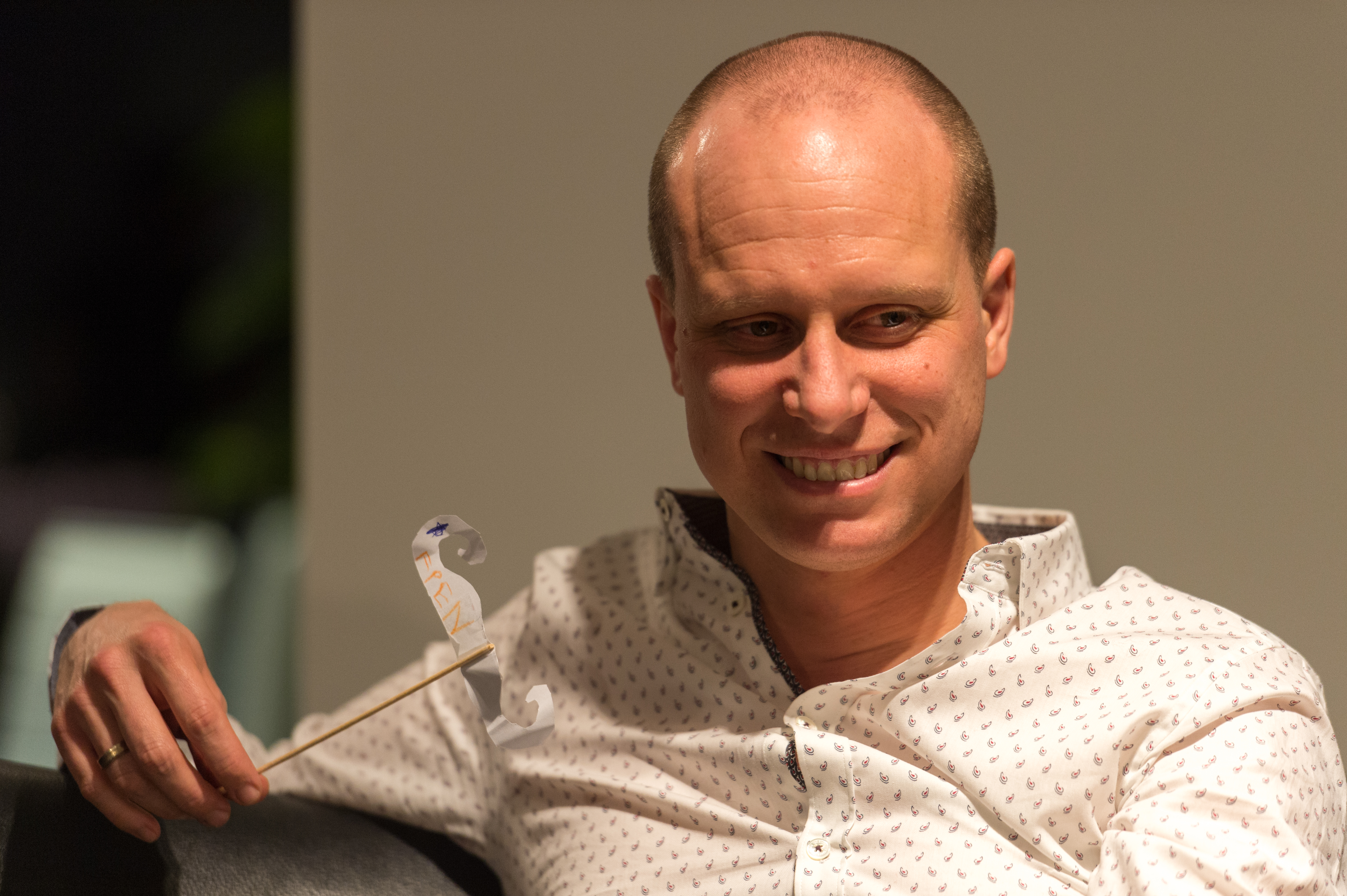 A man in a white patterned shirt smiles while holding a small decorative prop on a stick.