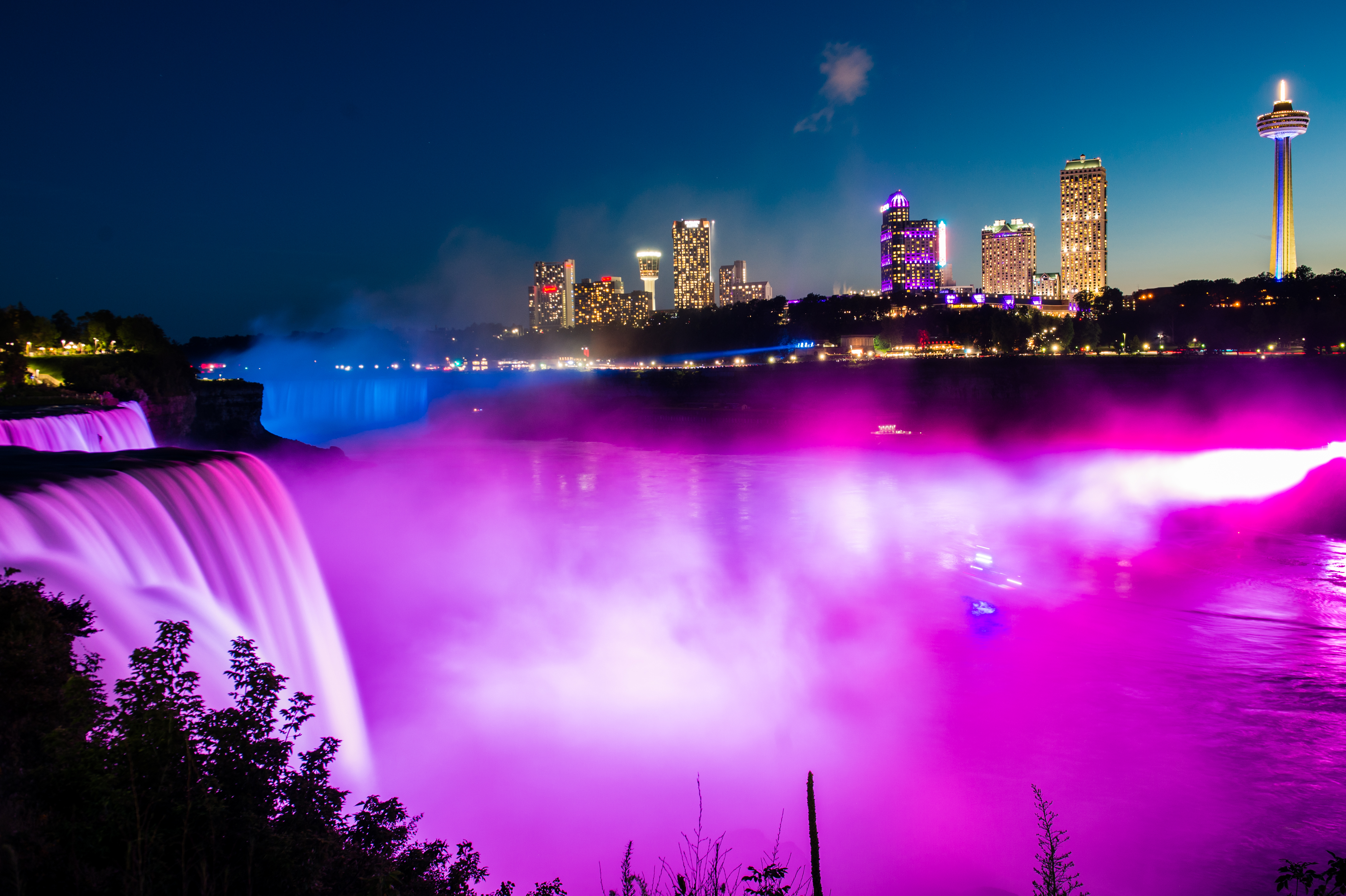 Niagara Falls glows with pink and purple lights at night, with a city skyline in the background.