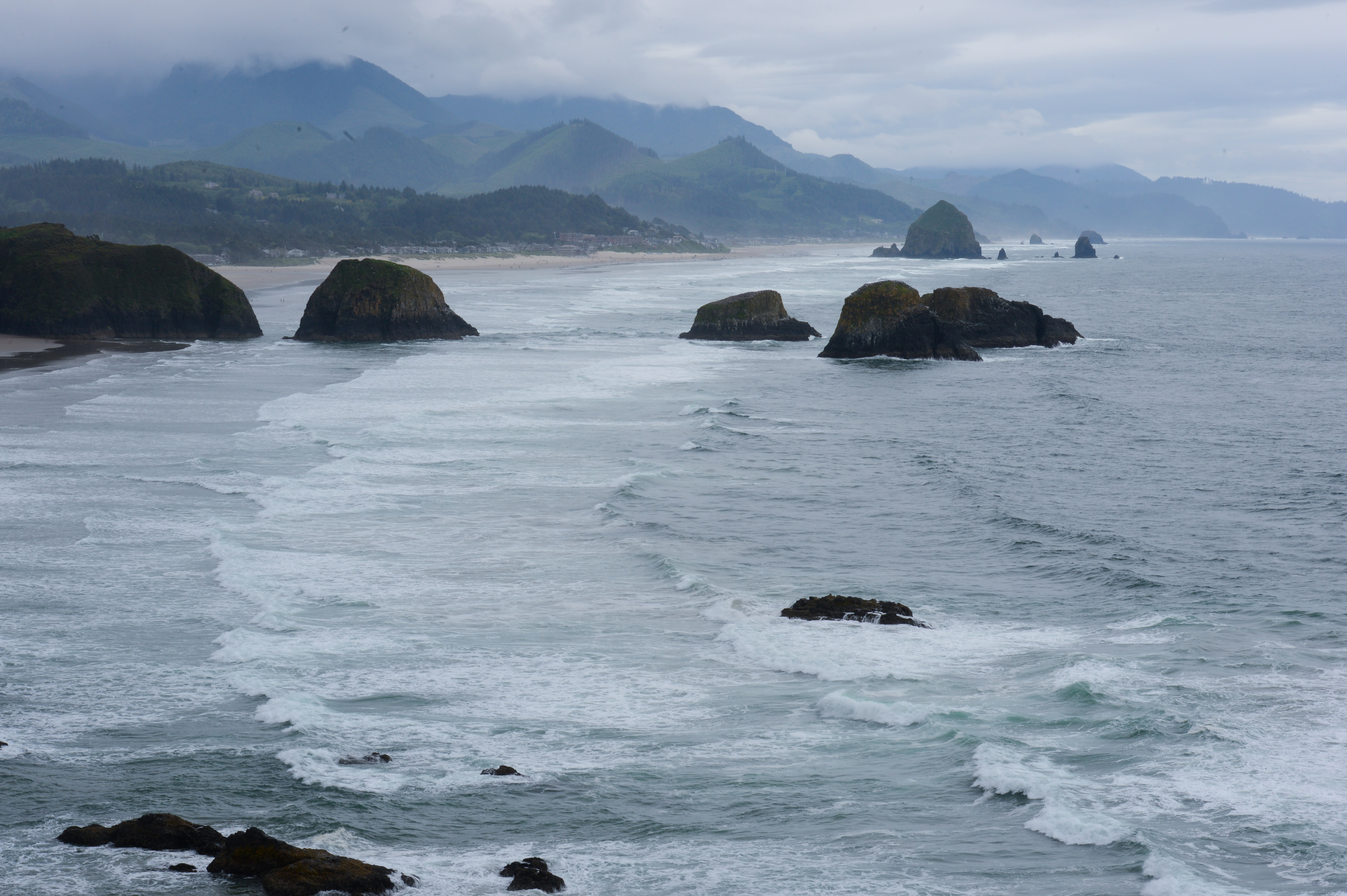 Waves crash against rocky sea stacks along the shoreline at Cannon Beach, with mountains and clouds in the background.