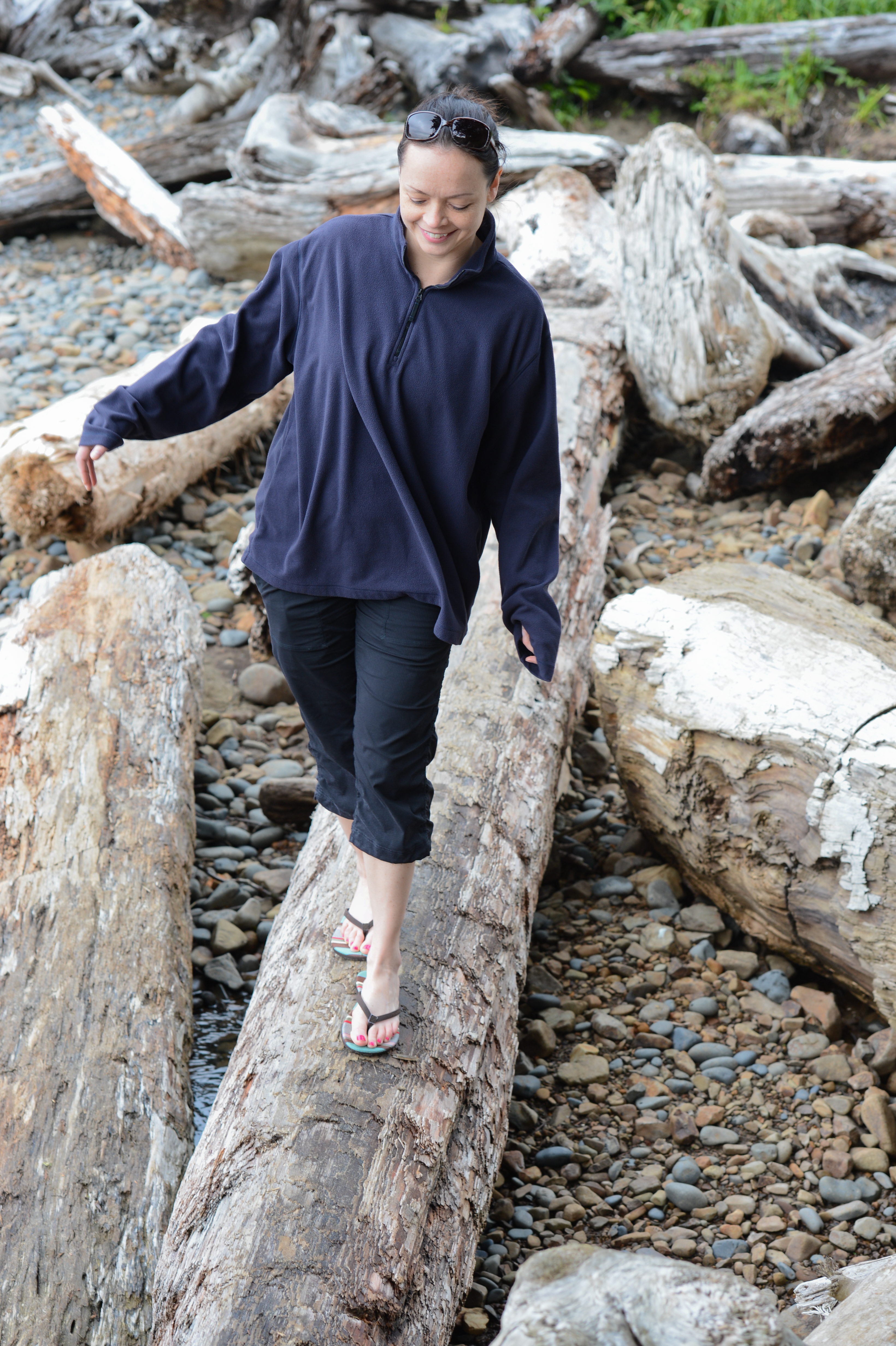 A woman in a dark jacket walks carefully on a large driftwood log at a rocky beach.