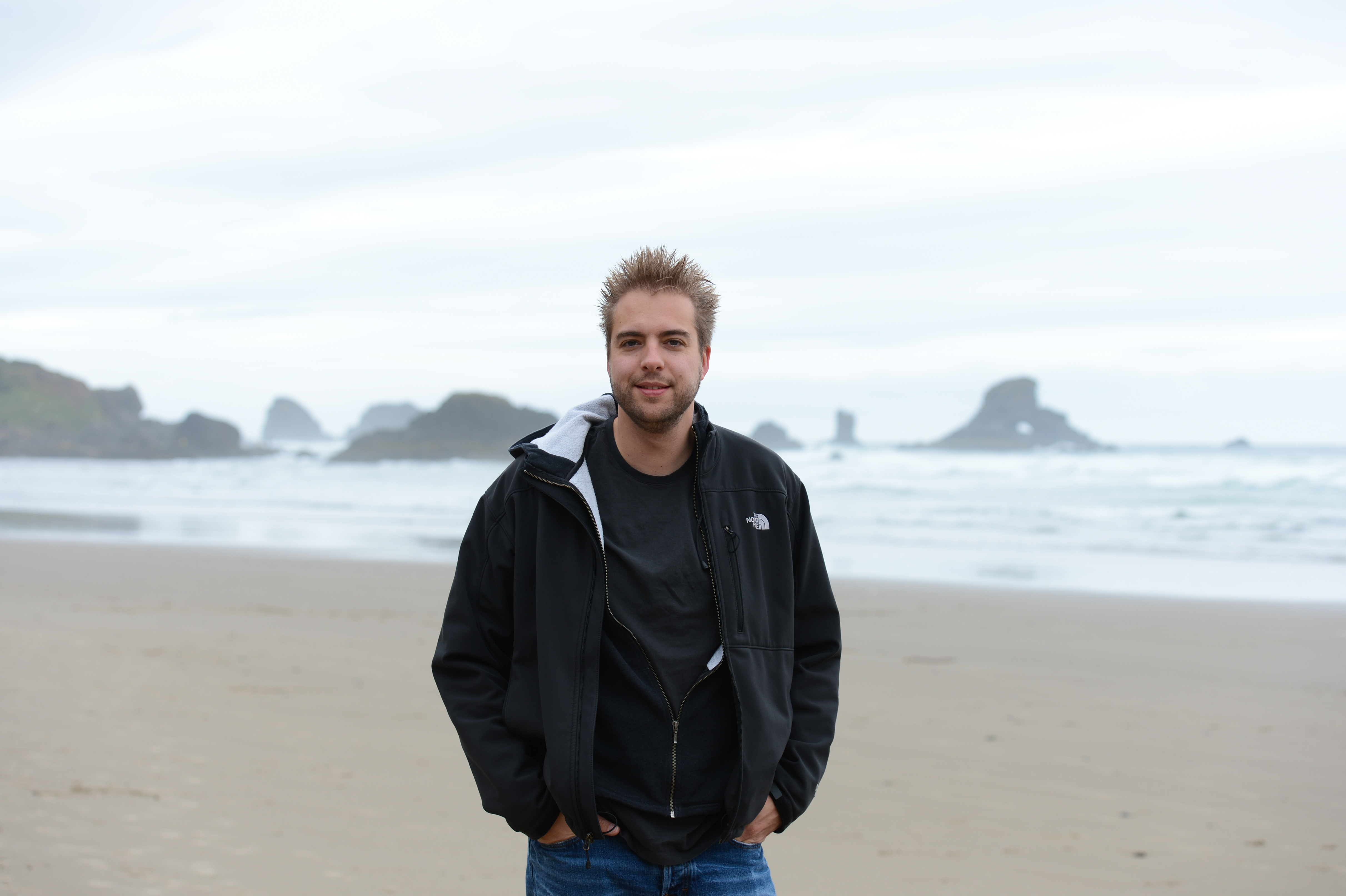 A man in a black jacket stands on a sandy beach with ocean waves and rock formations in the background.