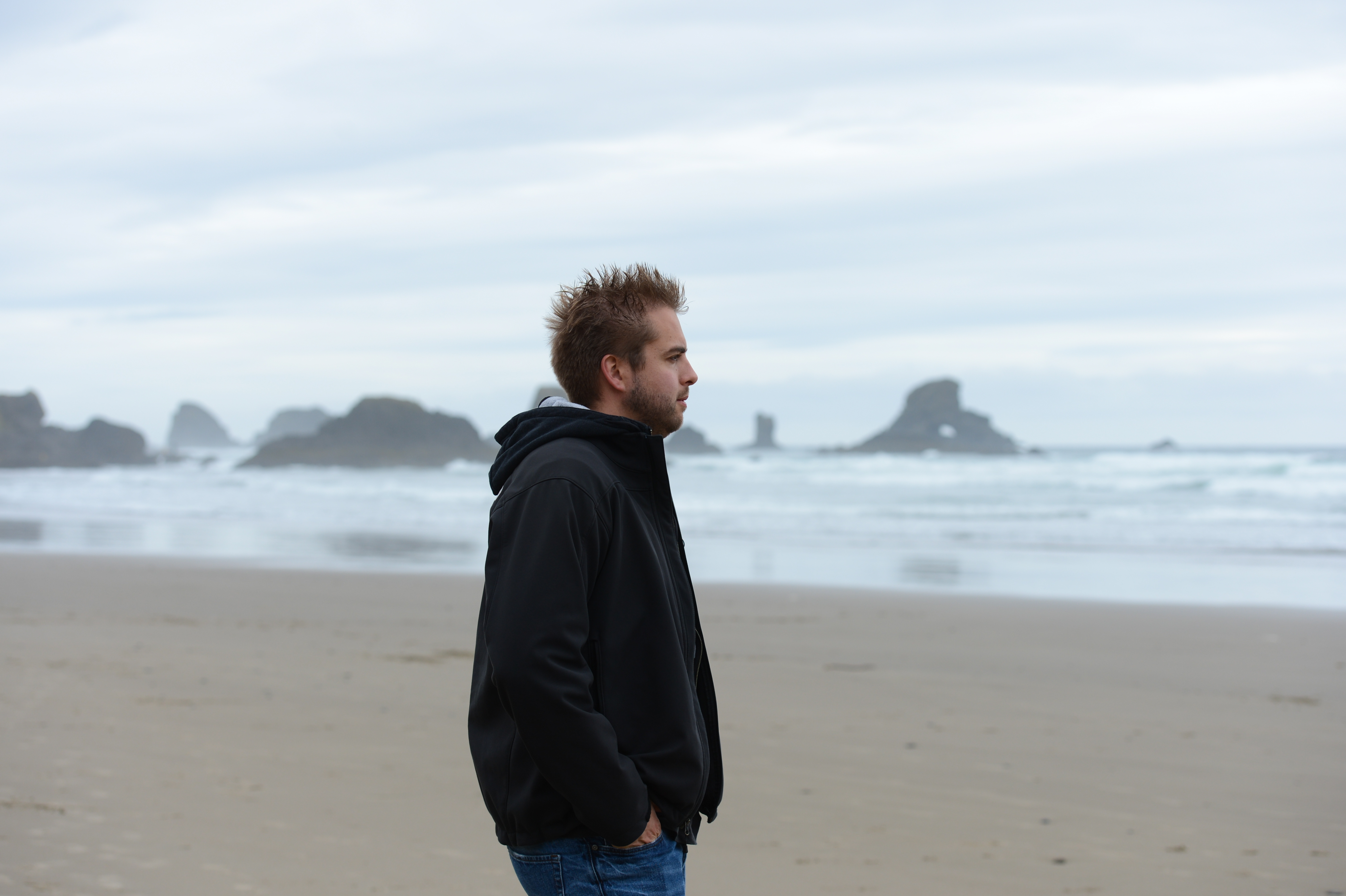 A person in a black jacket stands on a sandy beach, looking toward the ocean with rock formations in view.