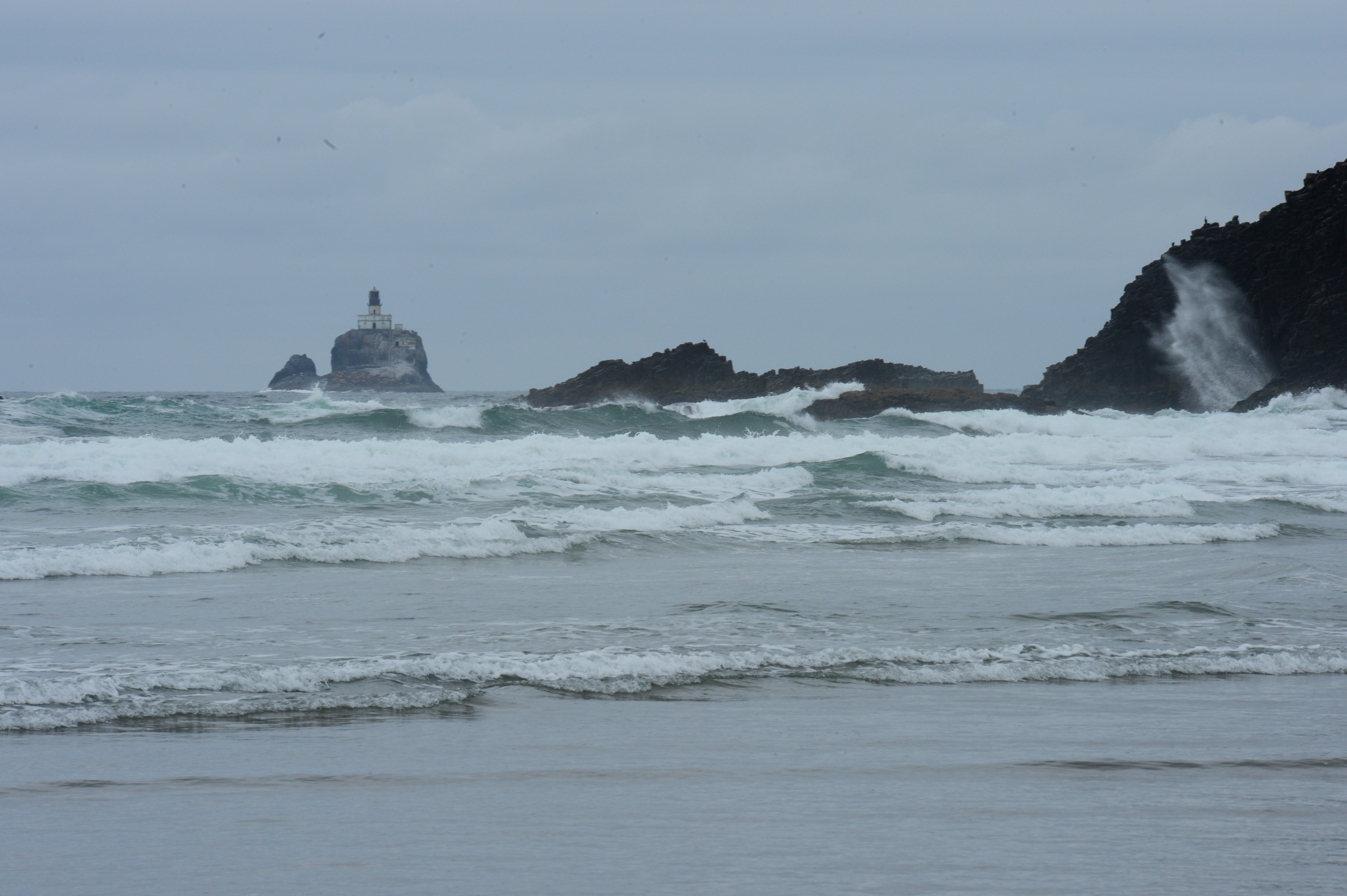 Waves crash against rocky cliffs with a lighthouse on a distant island under a cloudy sky.