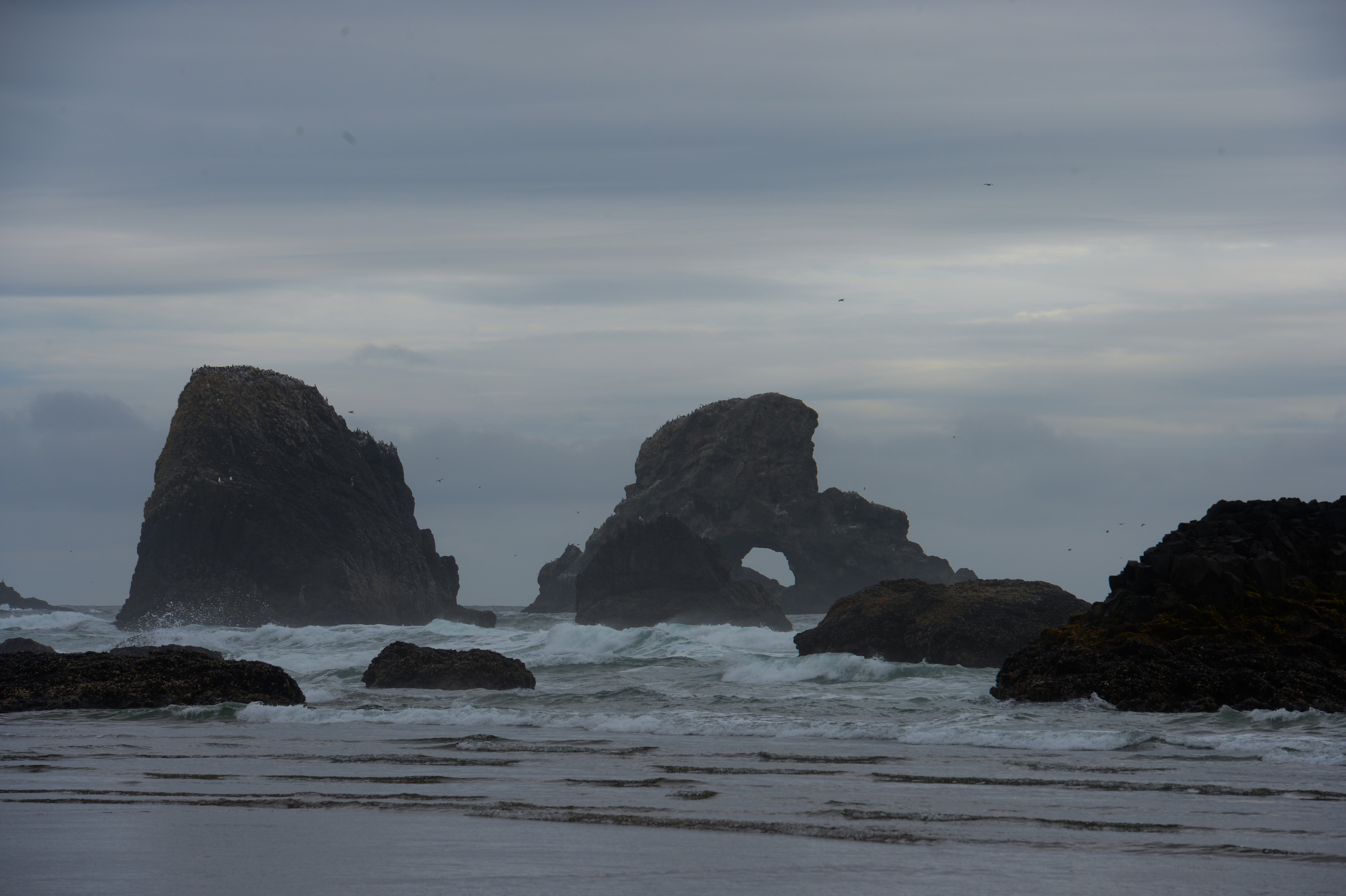 Rock formations rise from the ocean as waves crash against them under a cloudy sky at Cannon Beach.