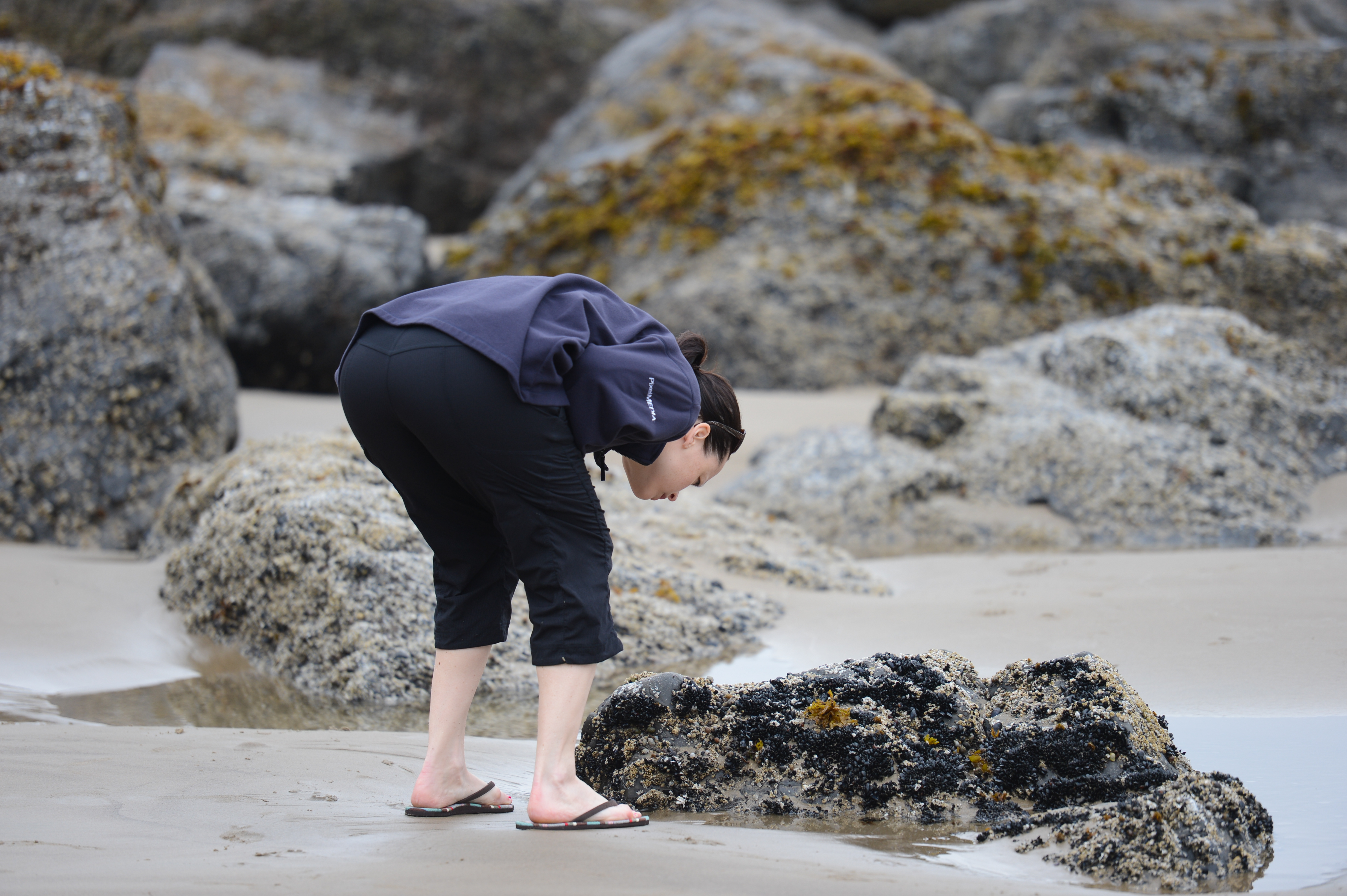 A person in a dark jacket and flip-flops bends down to examine a rock on the beach.