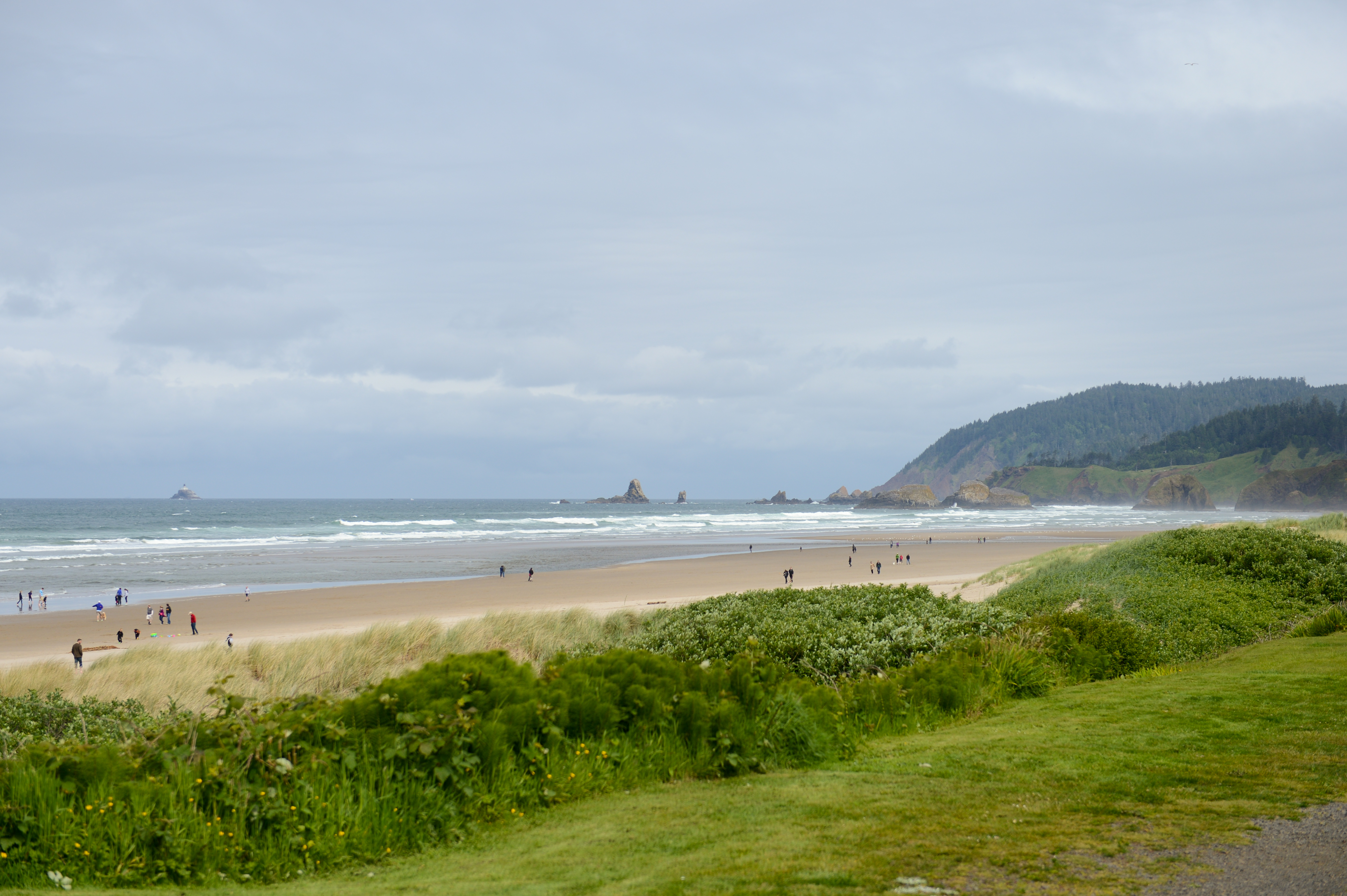 People walking along a wide sandy beach with ocean waves, distant rock formations, and green hills in the background.