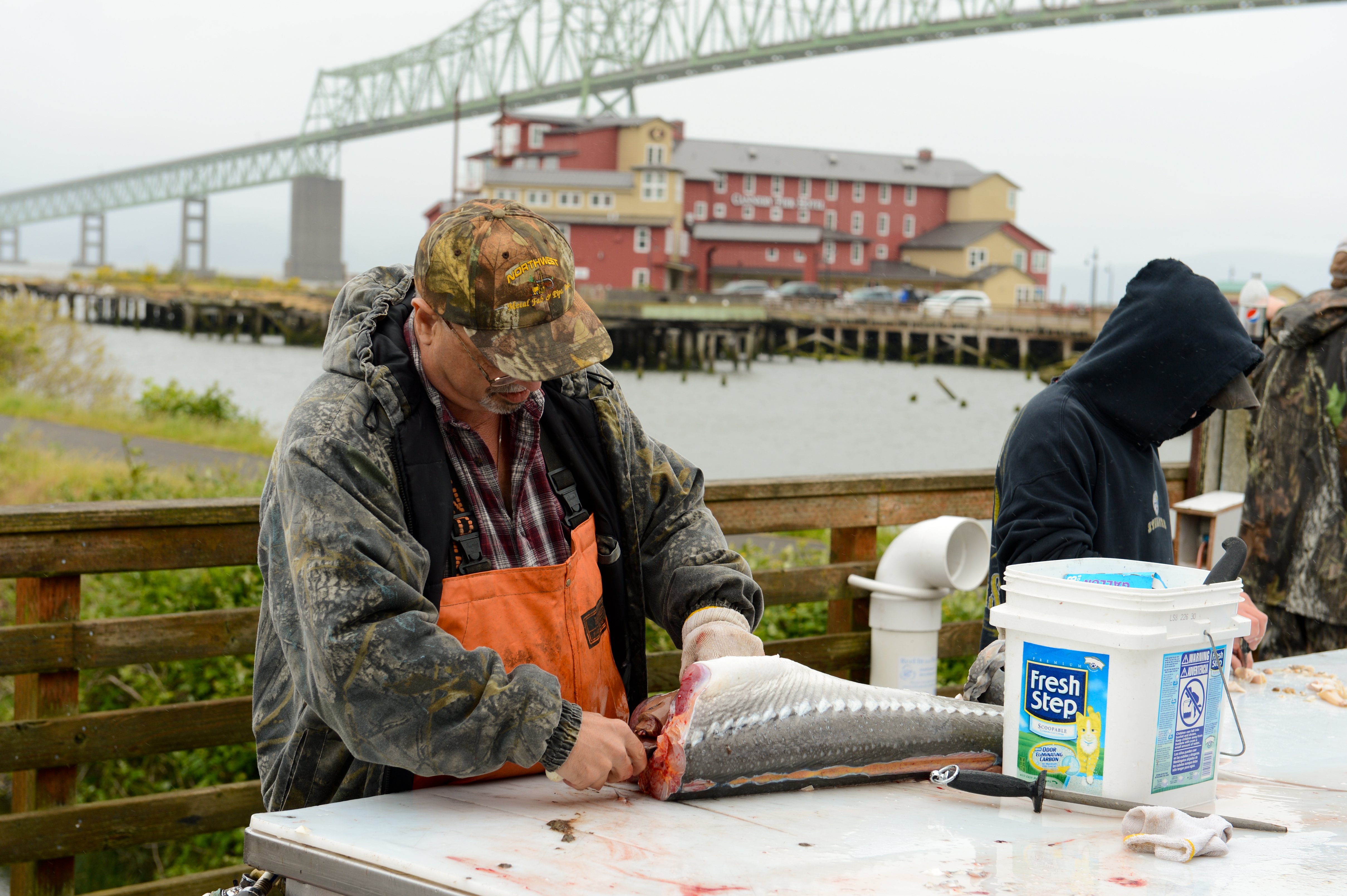 A man in a camouflage jacket and orange overalls fillets a large fish on an outdoor table.