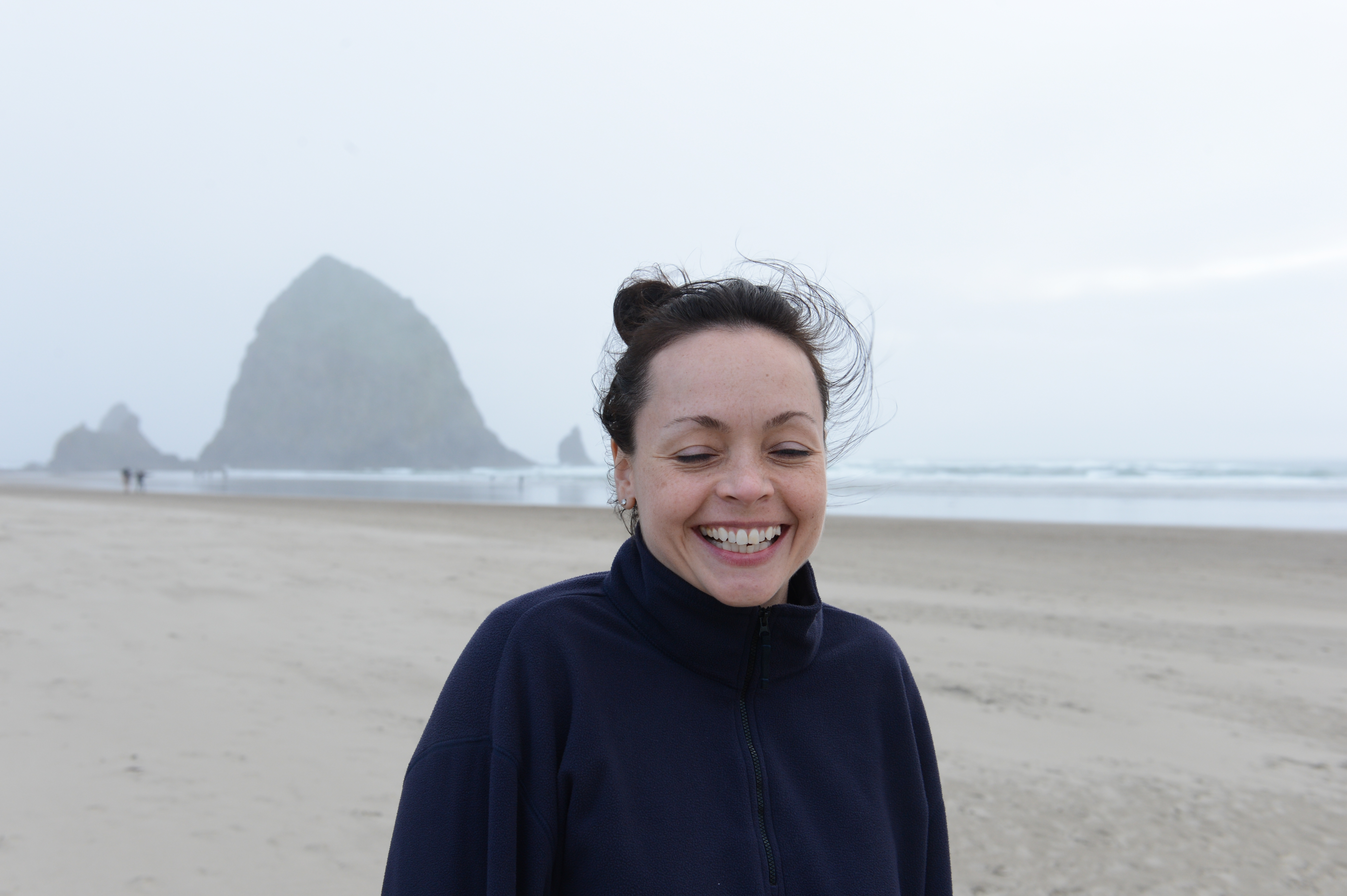 A smiling woman in a dark jacket stands on a sandy beach with large rock formations in the background.