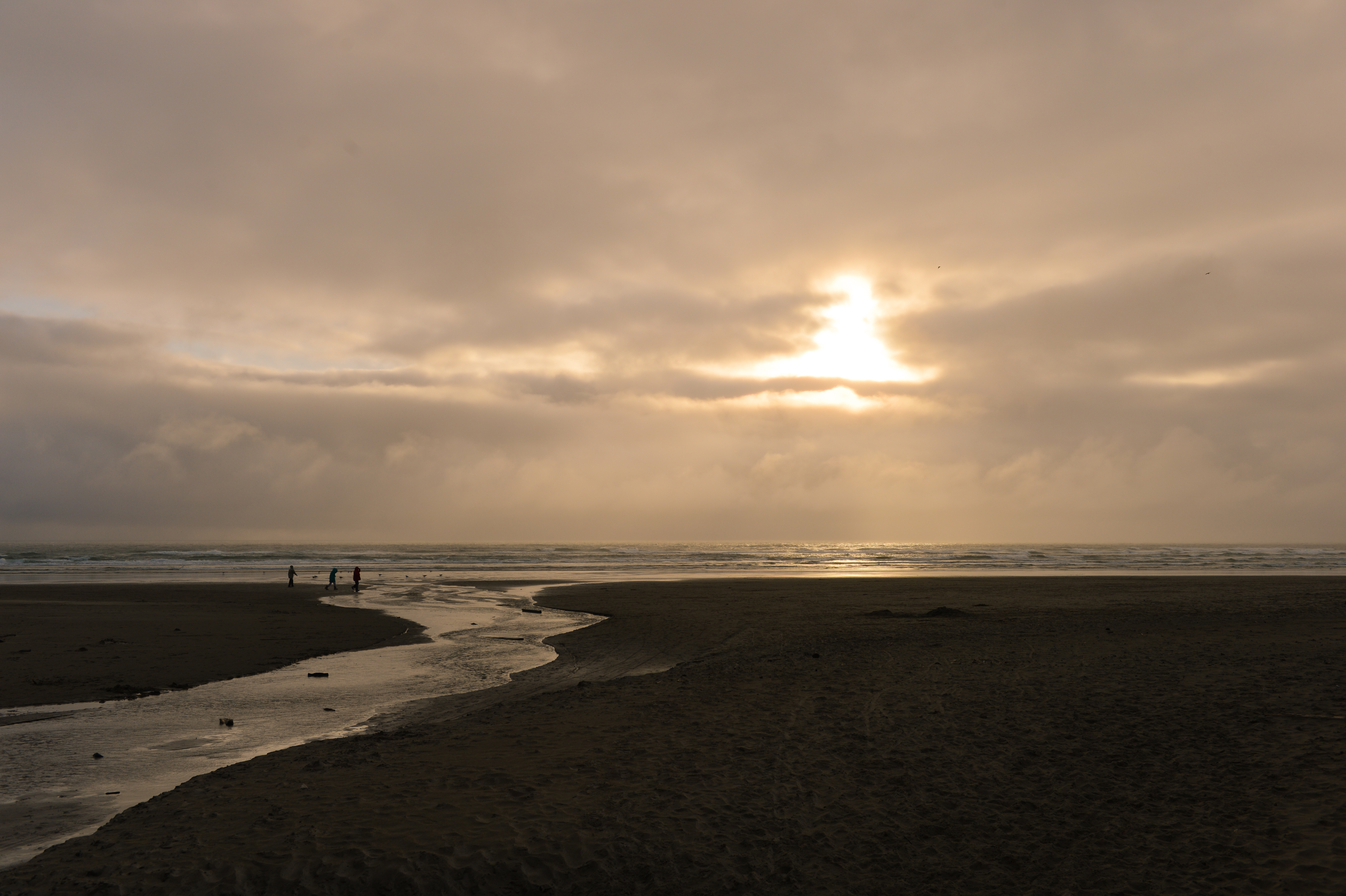 A few people walk along the shoreline at Cannon Beach under a cloudy sky with the sun shining through.