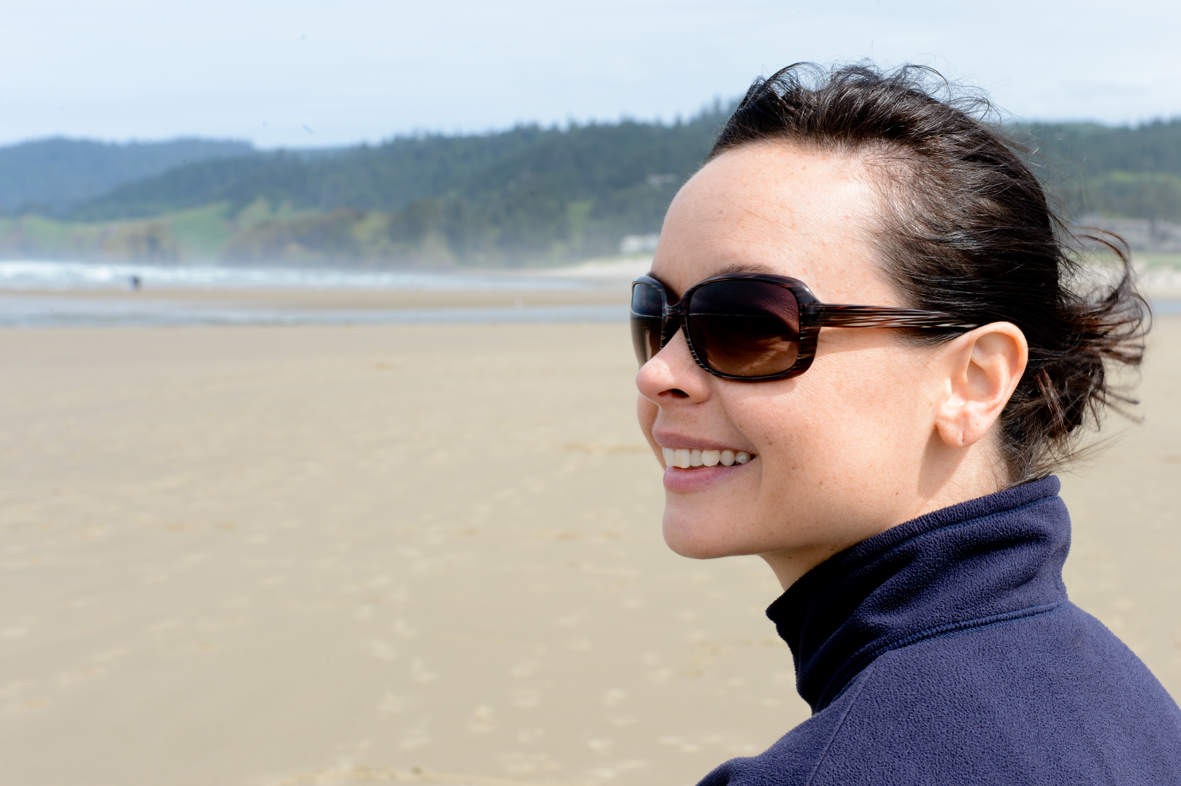 A woman wearing sunglasses and a dark jacket smiles while standing on a sandy beach with ocean waves behind her.