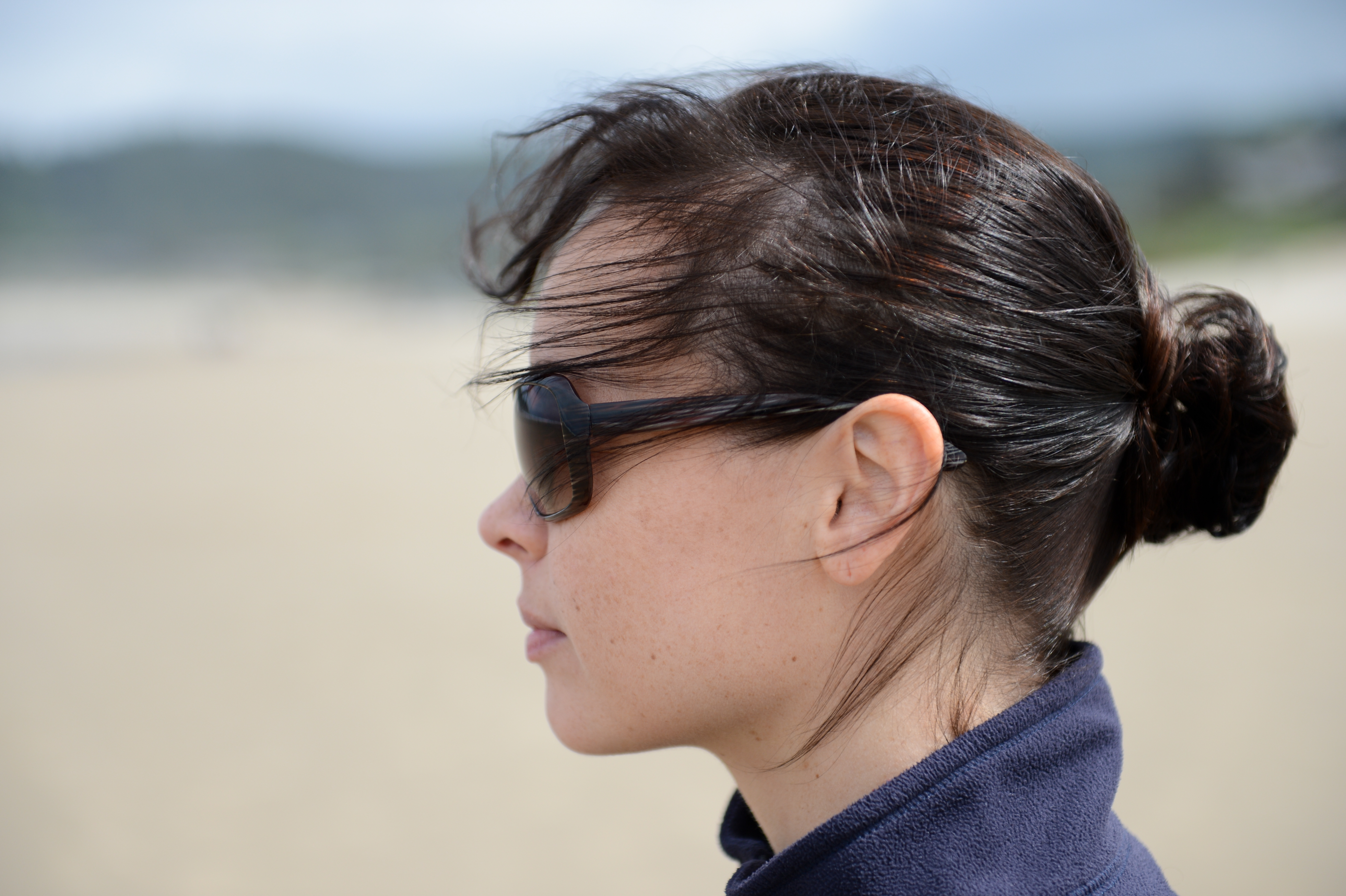 A woman wearing sunglasses and a dark jacket looks into the distance on a sandy beach.