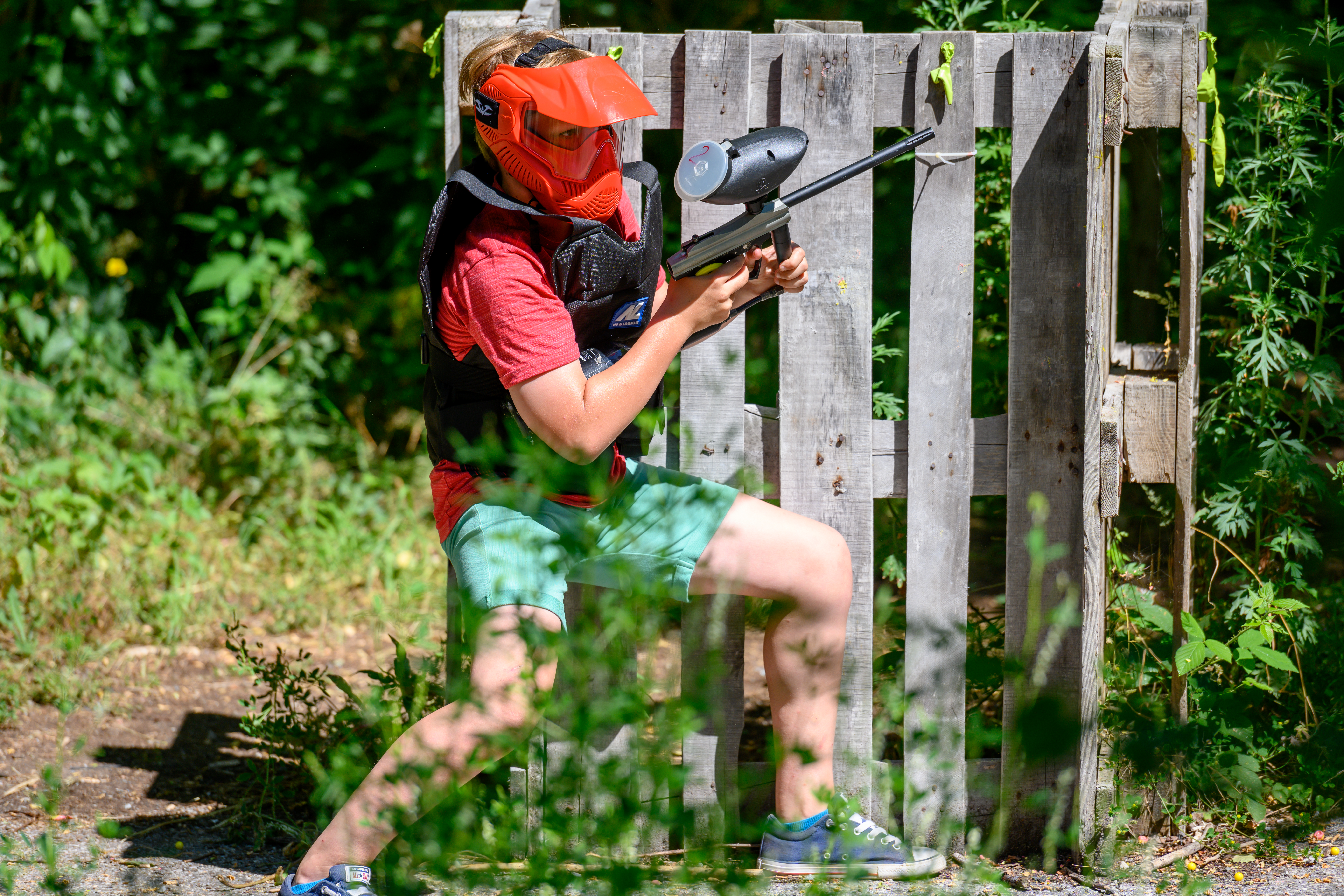 A person wearing a red mask and vest crouches behind a wooden barrier, aiming a paintball gun.