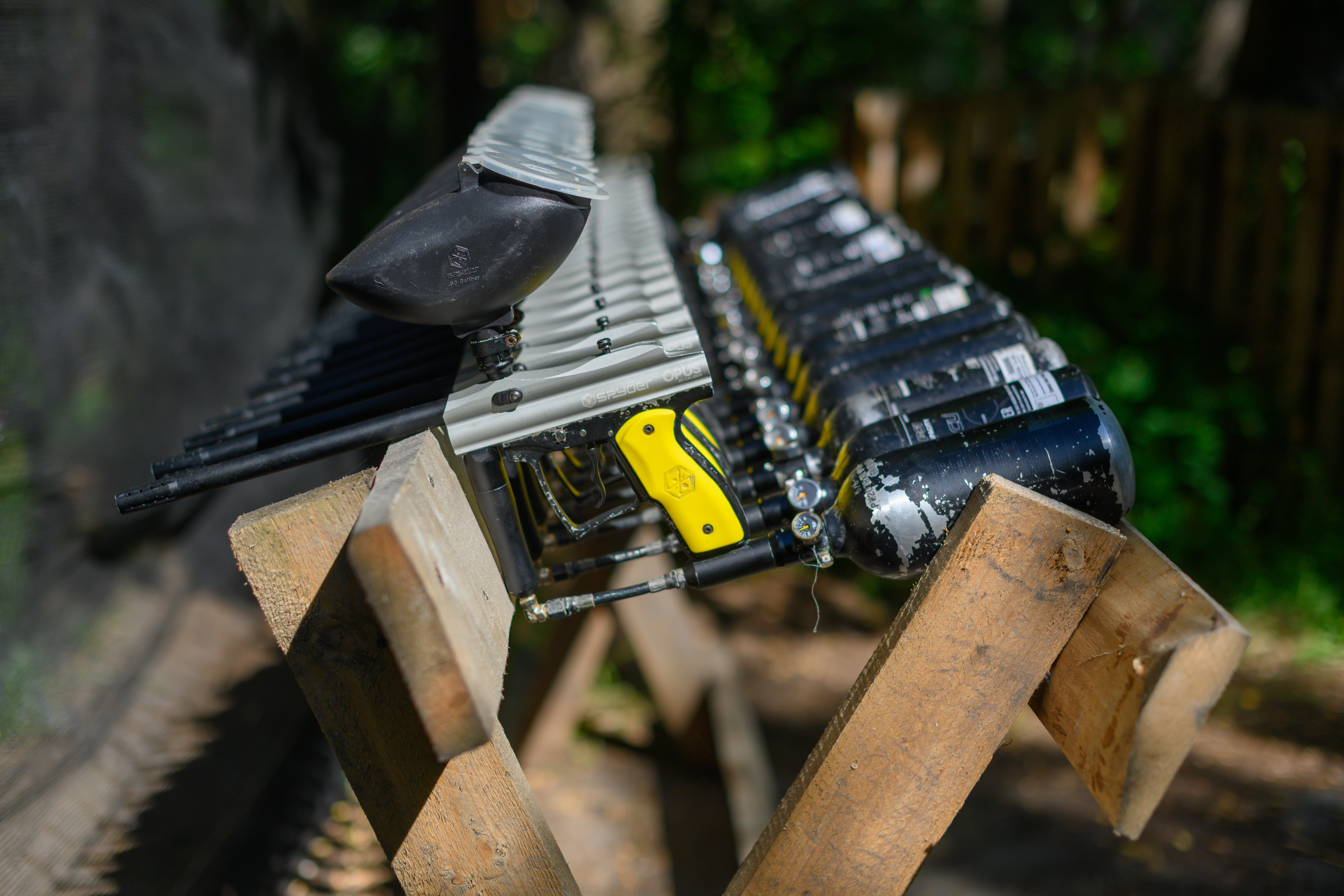 A row of paintball guns with air tanks lined up on a wooden stand outdoors.
