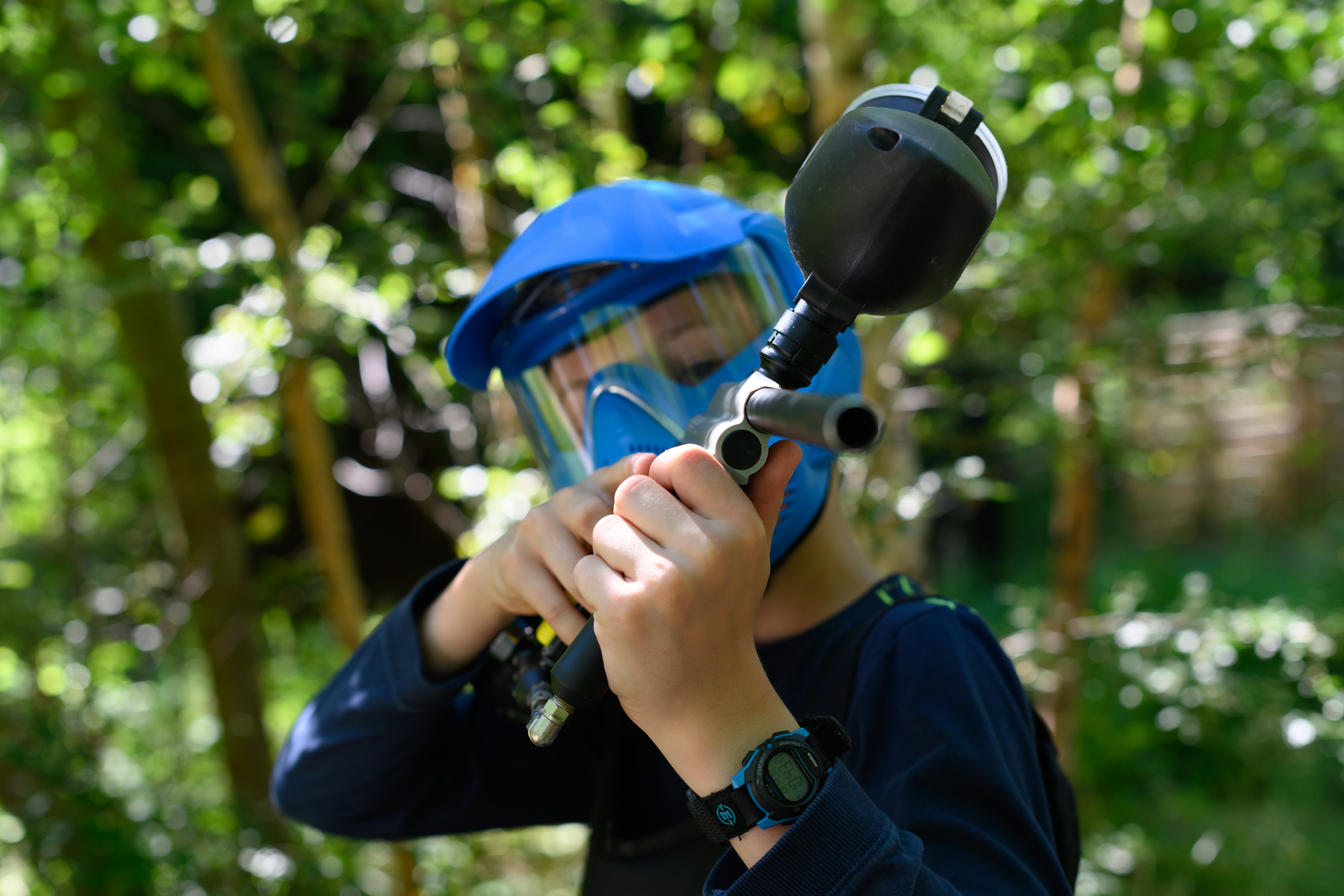 A person wearing a blue helmet and protective mask aims a paintball gun in an outdoor setting.