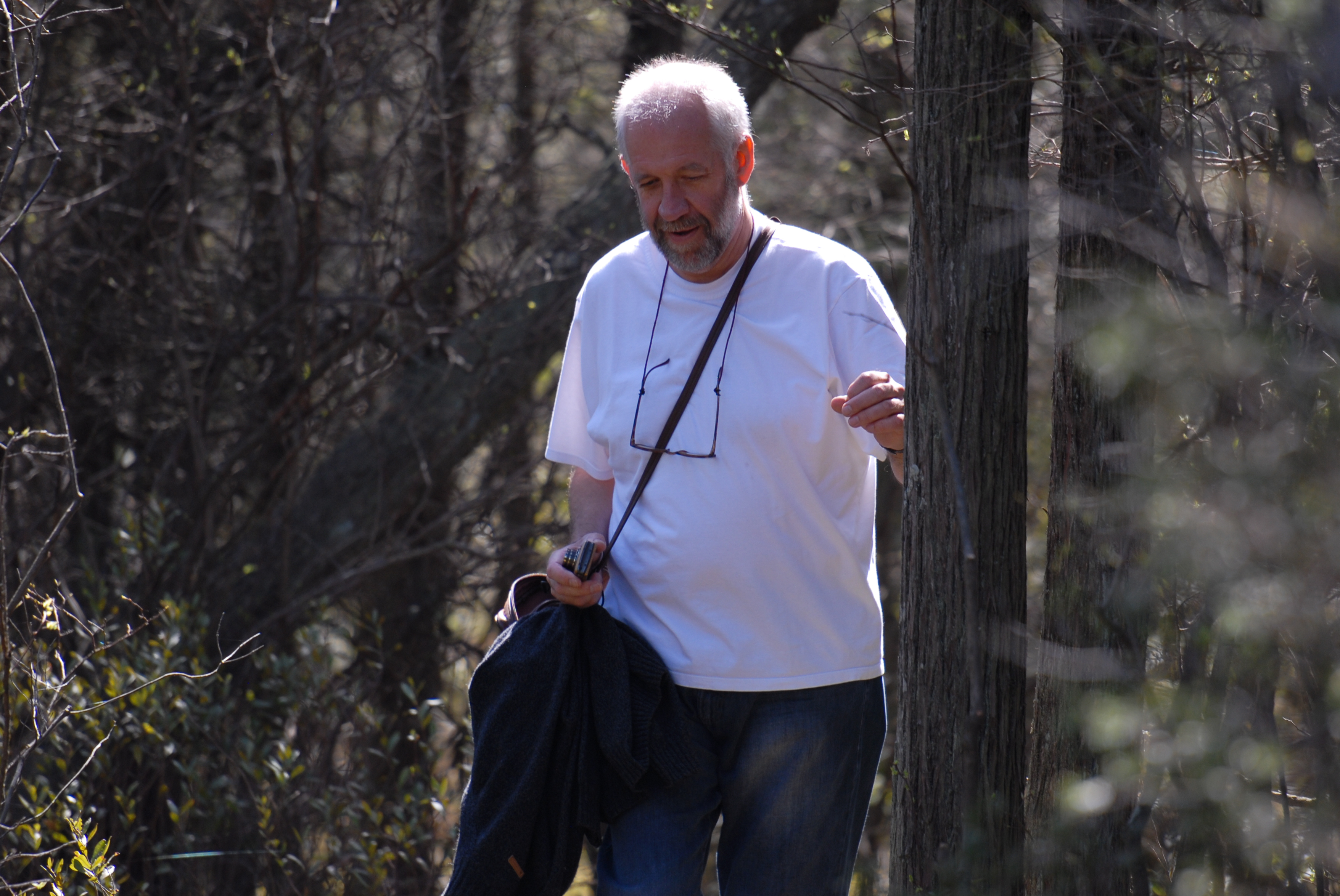 A man in a white shirt walks along a wooded trail, holding a jacket and a small object in his hand.