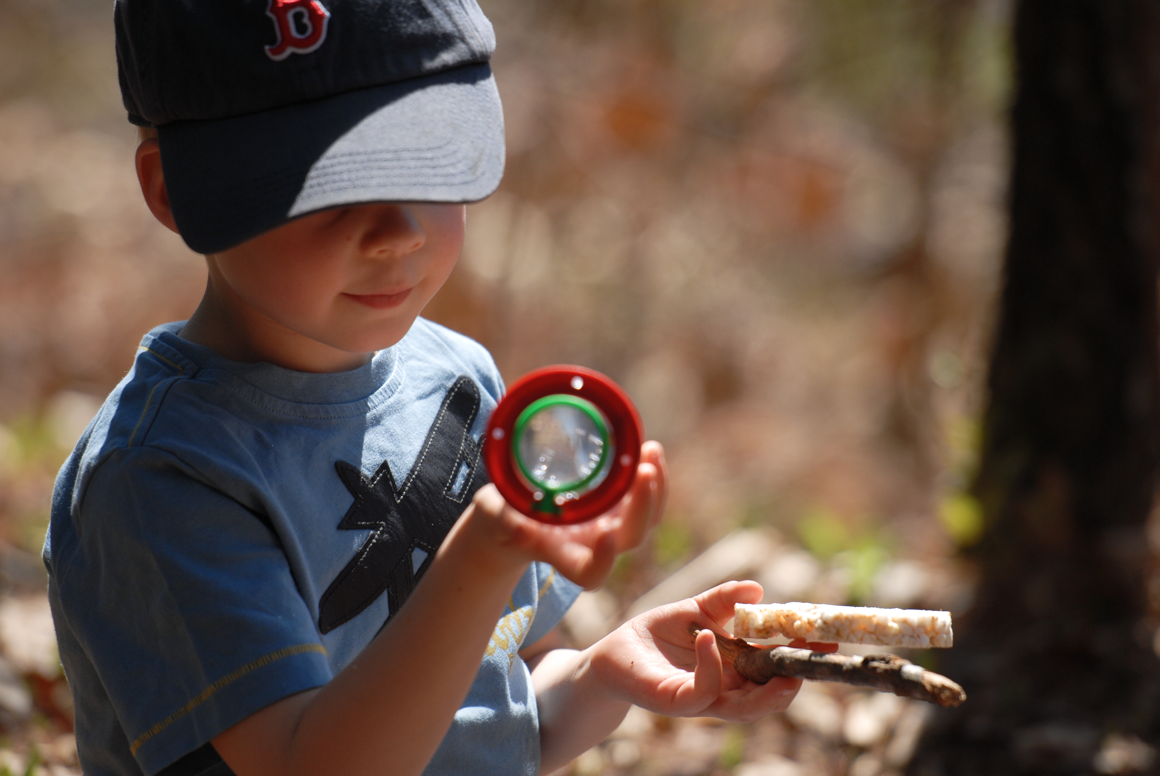 A young child wearing a baseball cap examines an object through a magnifying glass while holding a stick and snack.
