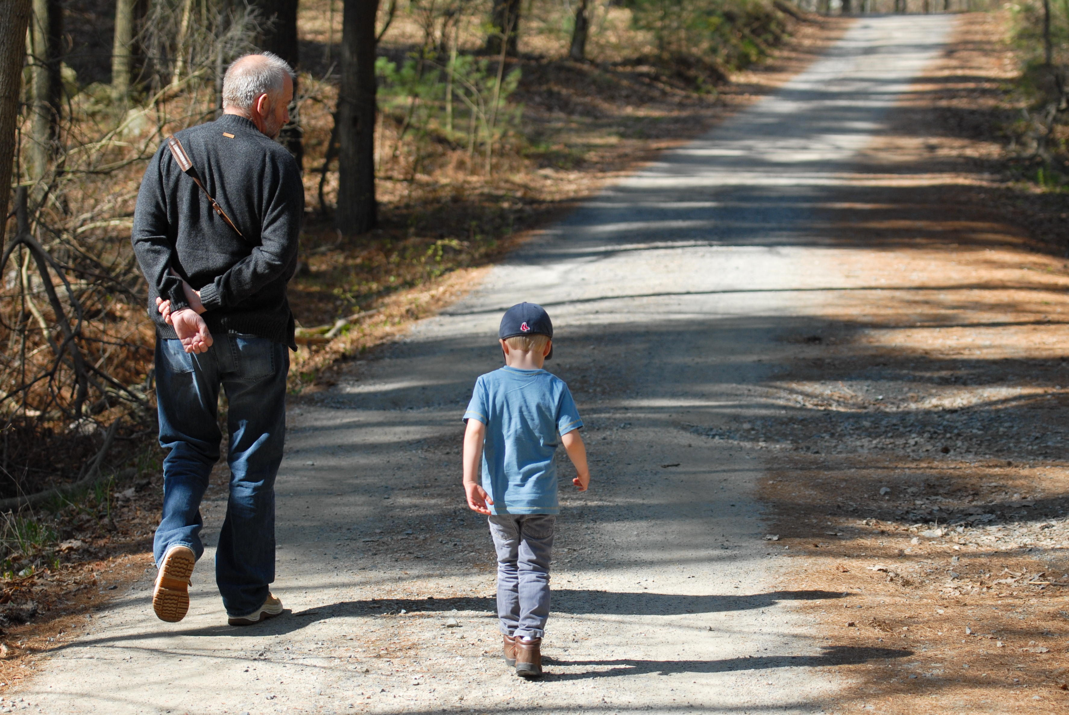 An older man and a young child walk along a dirt trail surrounded by trees.