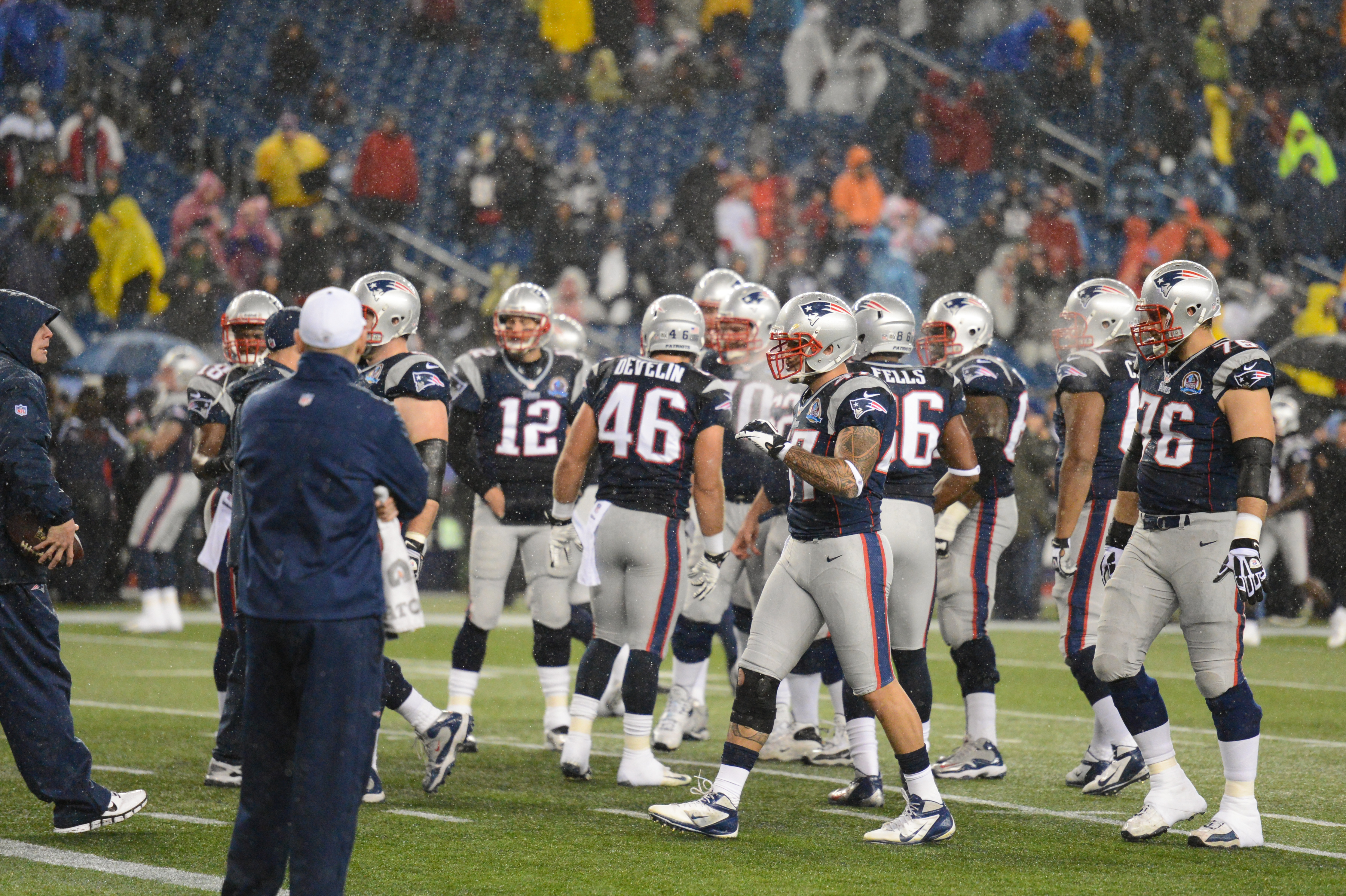 New England Patriots players huddle on the field during a rainy game, wearing helmets and uniforms.