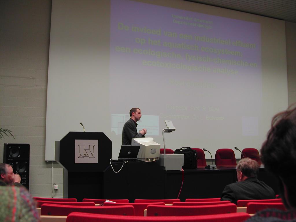 A man in a suit stands at a podium, presenting his PhD defense in a lecture hall.