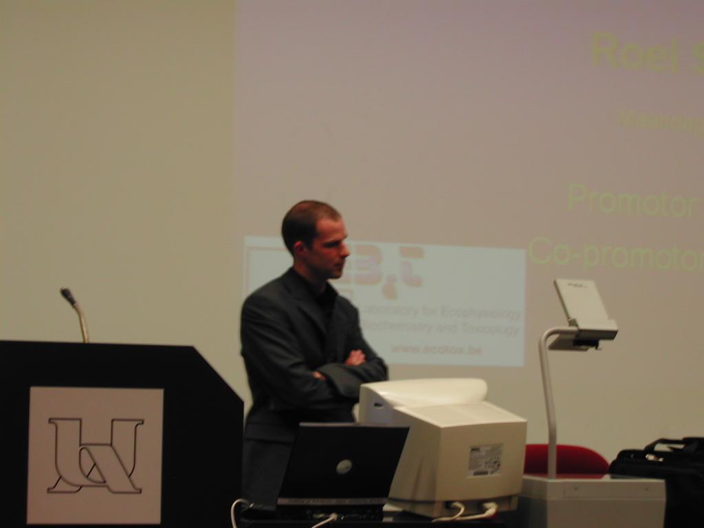 A man in a suit stands in front of a projector screen during a PhD defense presentation.