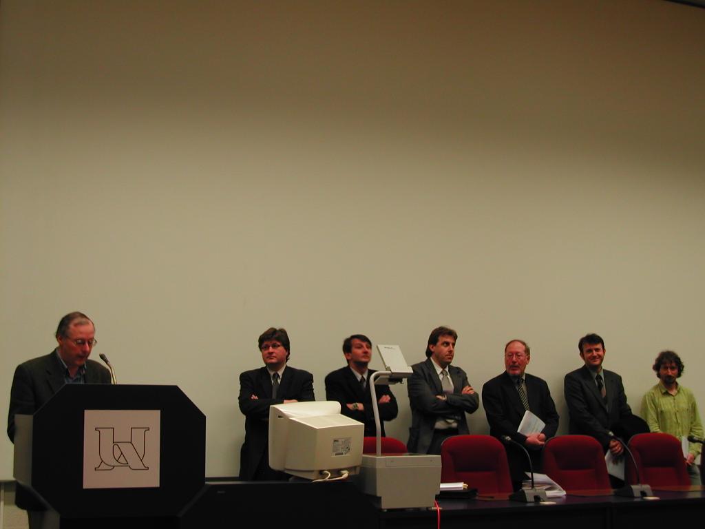 A man speaks at a podium while six others stand behind him during a PhD defense event.